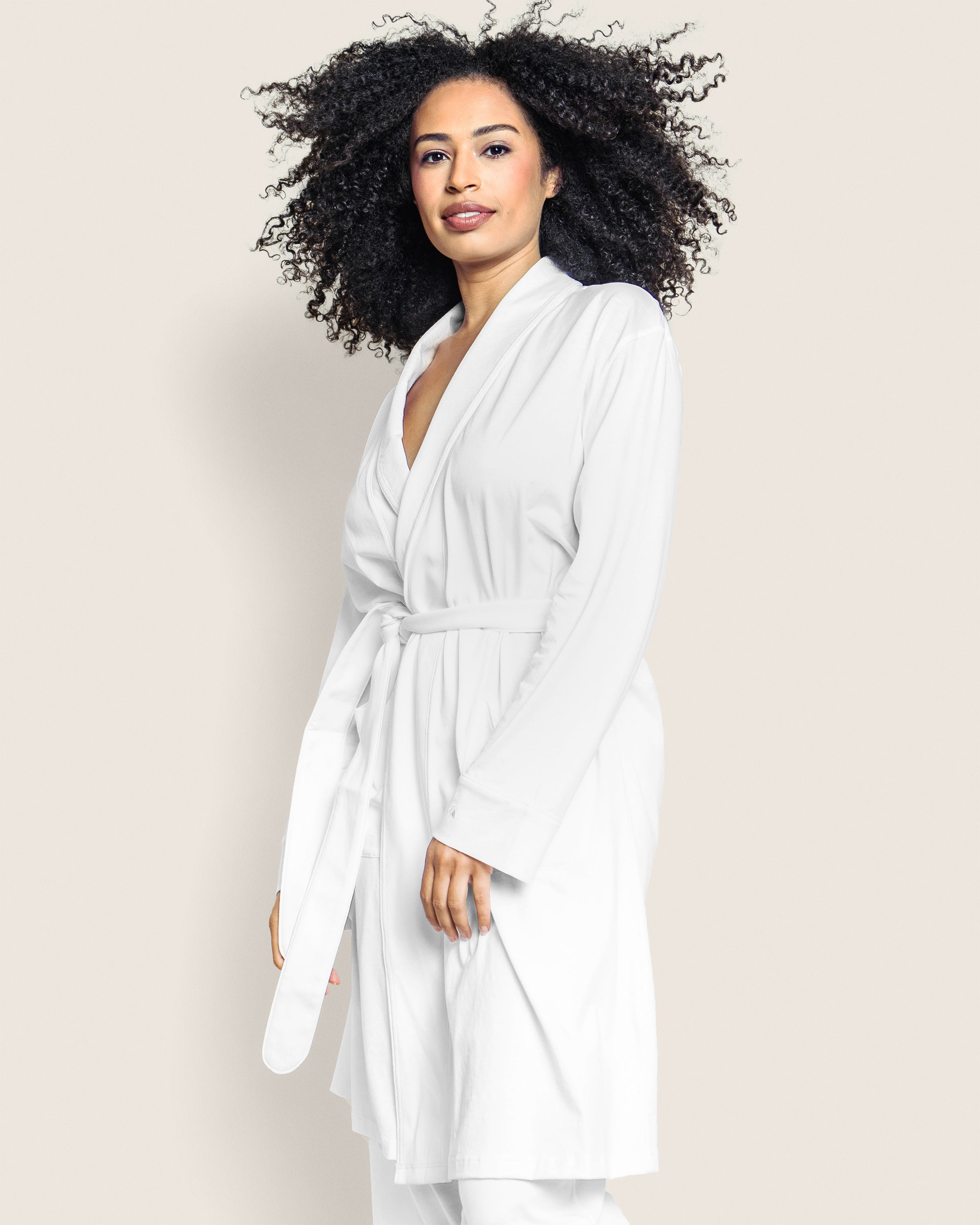 A person with curly hair wears a luxurious Womens Pima Robe in White by Petite Plume, standing against a plain background and looking directly at the camera with a slight smile.