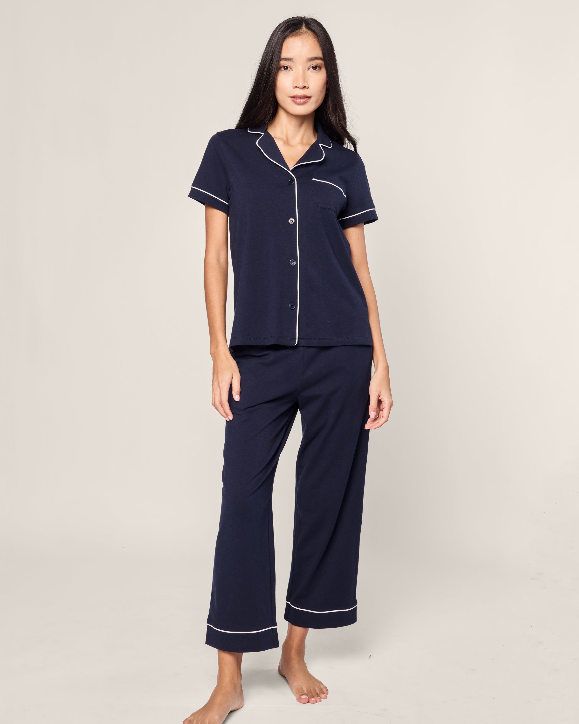 A person with long dark hair wears the Womens Pima Crop Set in Navy with White Piping by Petite Plume. The luxurious Peruvian Pima cotton set features a short-sleeved button-up shirt and long pants, and they stand barefoot against a plain, light-colored background.