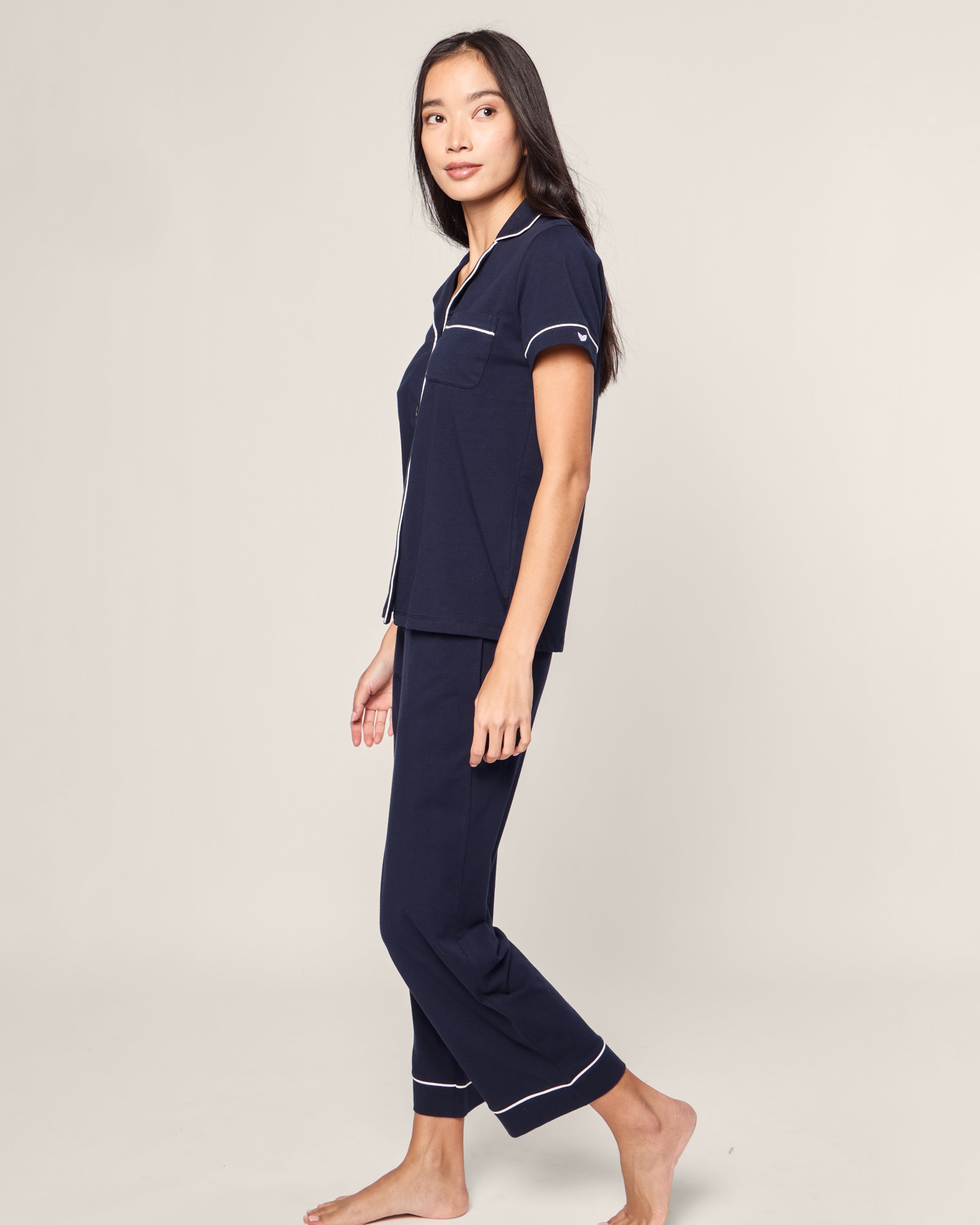 A person with long dark hair is wearing the Womens Pima Crop Set in Navy with White Piping by Petite Plume. They are barefoot, standing on a plain background, facing slightly to the side, and smiling.