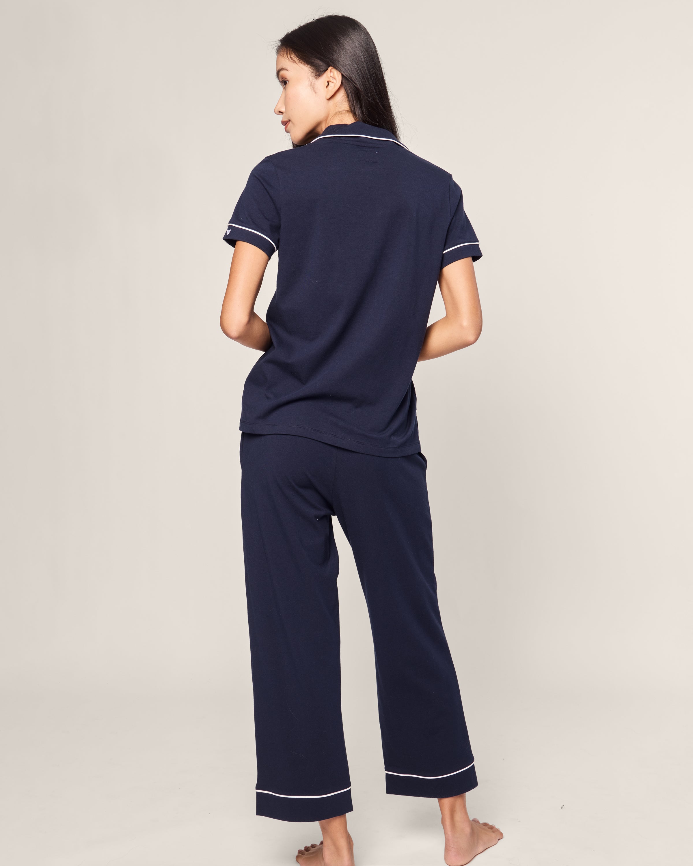 Barefoot and facing away, the person wears a luxurious Womens Pima Crop Set in Navy with White Piping by Petite Plume. Crafted from sumptuous Peruvian Pima cotton, the short-sleeve top and matching pants exude elegance against a plain, light-colored background.