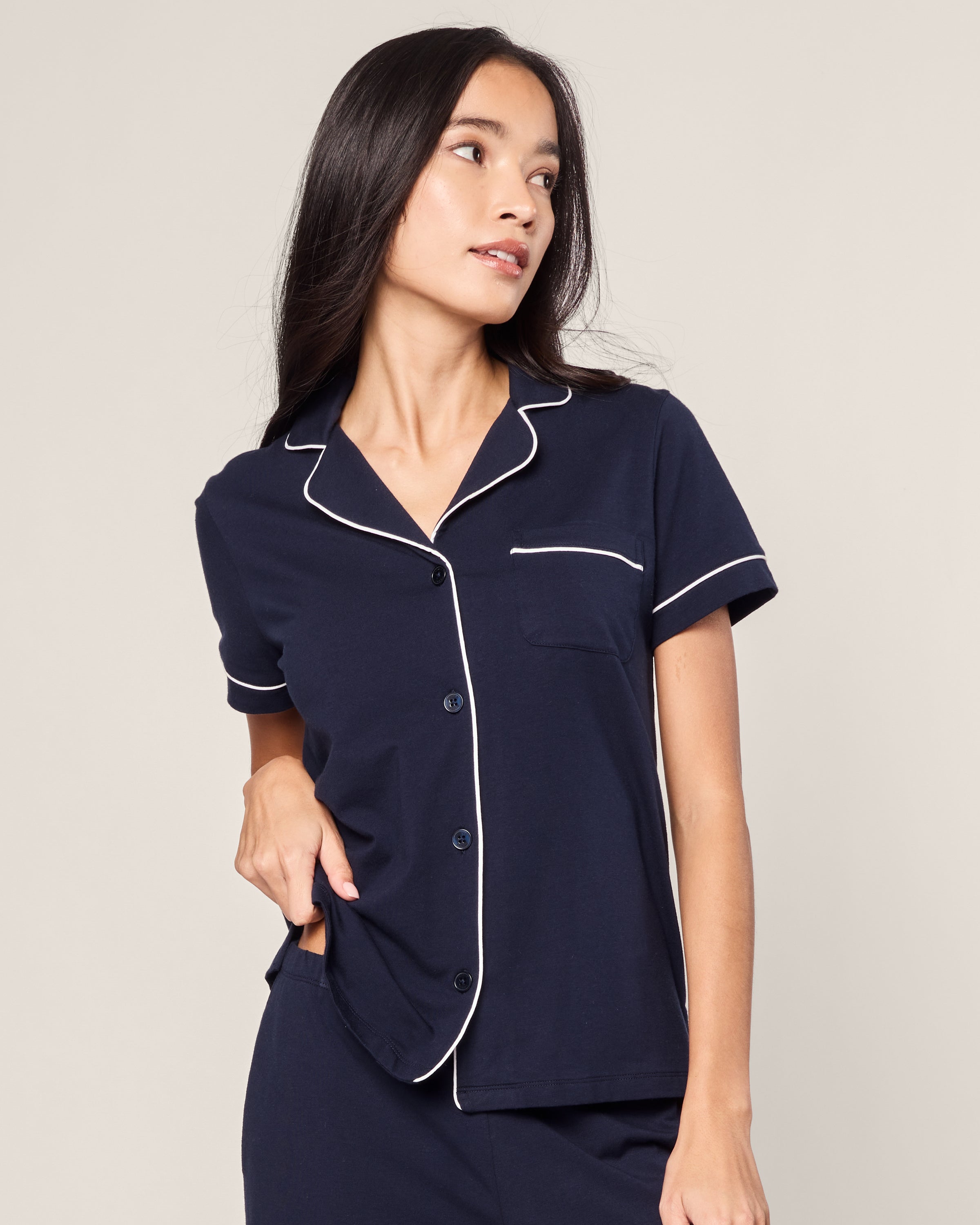 A person with long dark hair wears the Petite Plume Womens Pima Crop Set in Navy with White Piping, crafted from luxurious Peruvian Pima cotton. They glance to the side, gently lifting the shirts hem against a plain background.