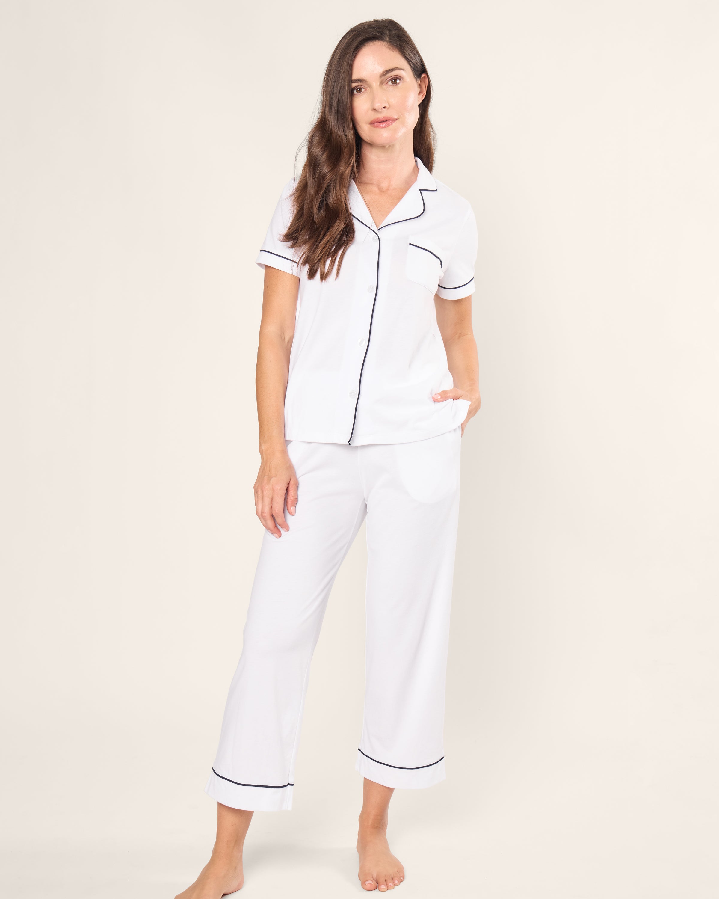 A woman stands barefoot in a Petite Plume Womens Pima Crop Set in white with navy piping. The luxurious Peruvian Pima cotton short-sleeve button-up top and pants offer a breathable feel. She has one hand in her pocket and displays a relaxed expression against a plain background.