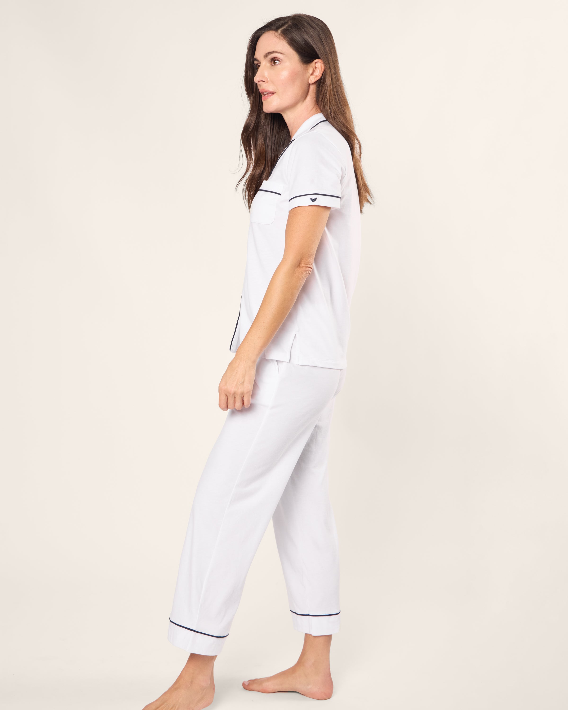 A woman with long brown hair stands sideways wearing the Womens Pima Crop Set in White with Navy Piping by Petite Plume, made from soft Peruvian Pima cotton. The jersey knit pajamas feature a small heart on the sleeve and provide a breathable feel against her skin, set against a neutral backdrop.