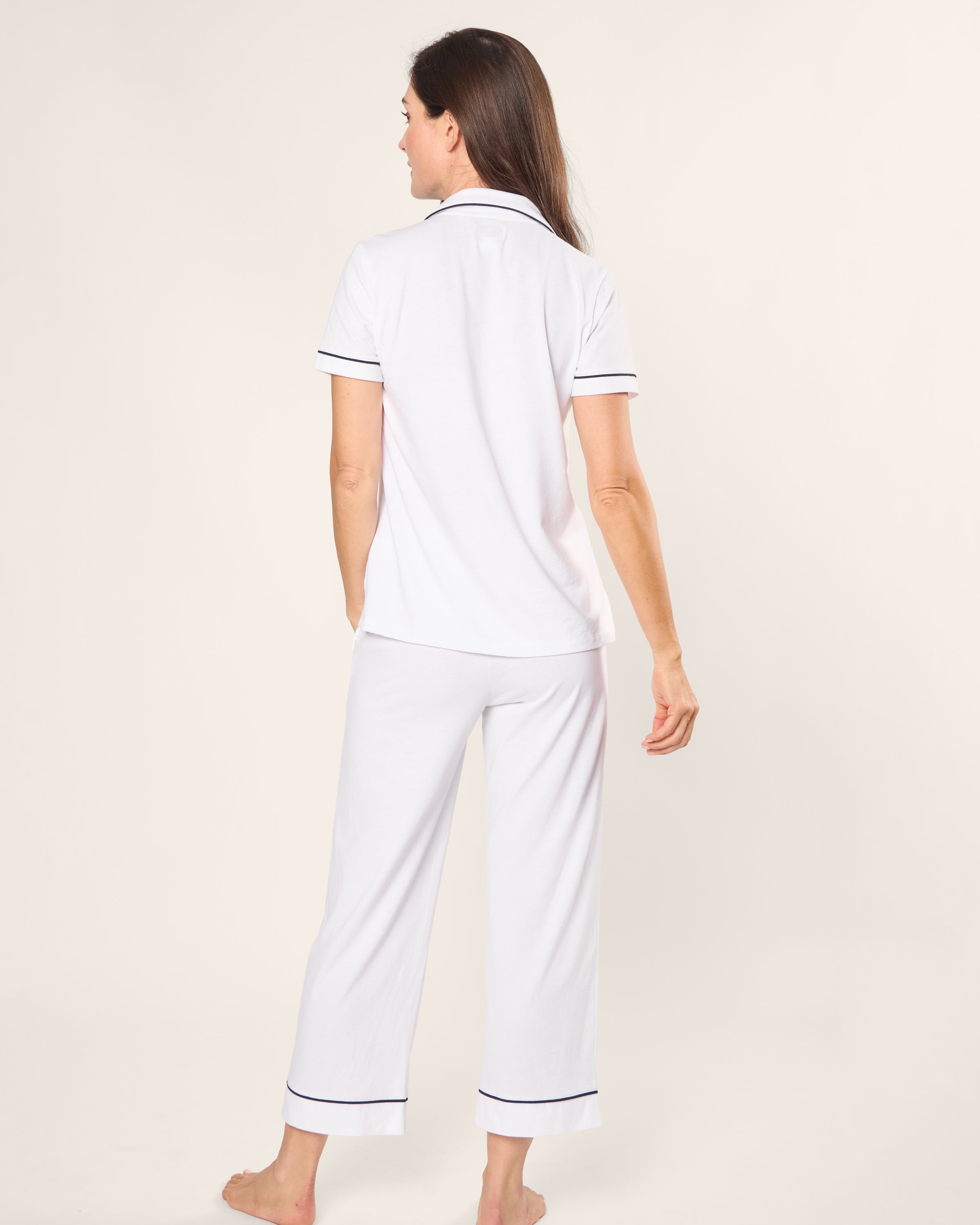 A person stands barefoot, facing away, in a Womens Pima Crop Set by Petite Plume. The white Peruvian Pima cotton pajama set features navy piping, and its collared top and cropped pants provide relaxed elegance against a plain light backdrop, enhancing the ensembles serene vibe.