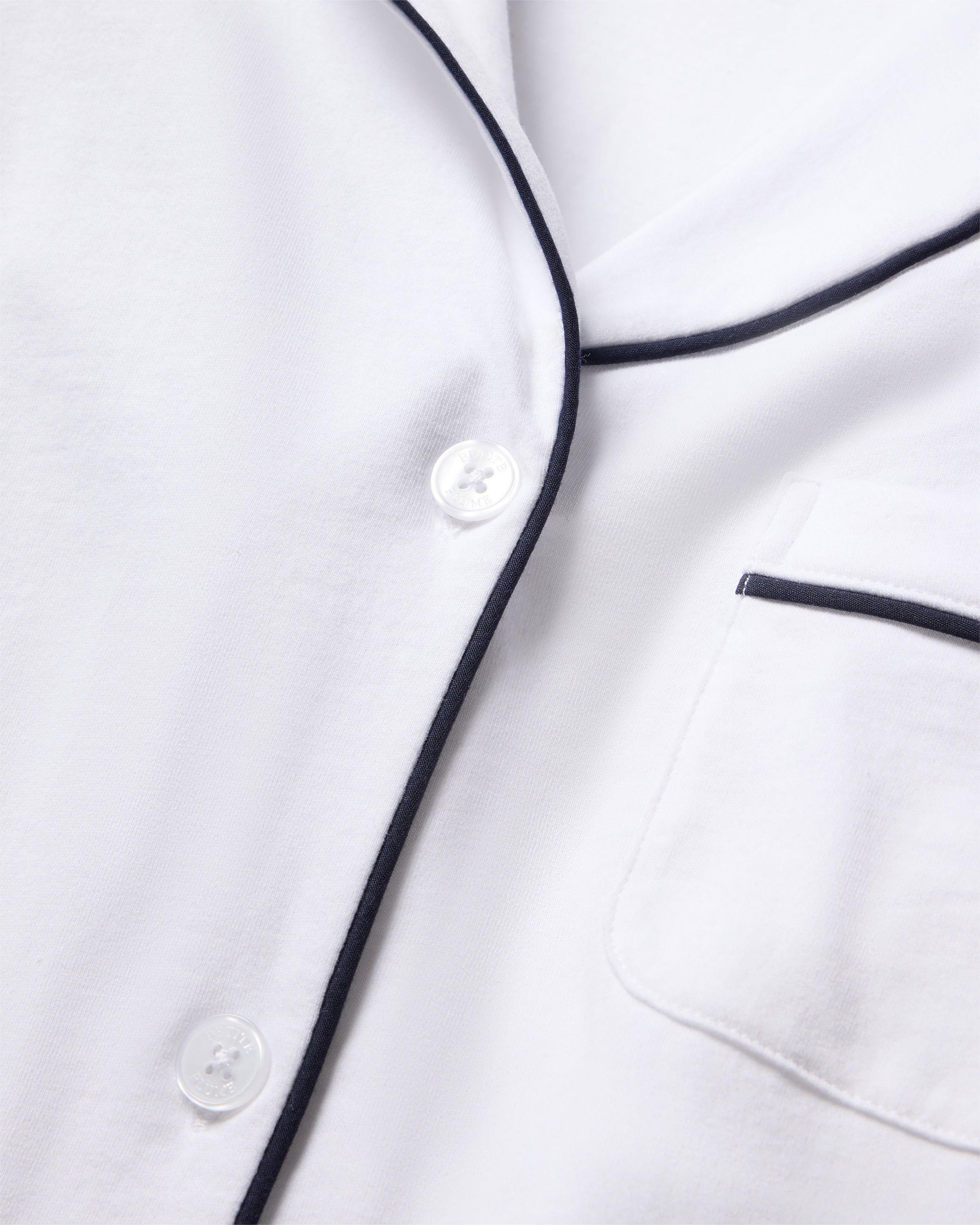 Close-up of Petite Plumes Womens Pima Crop Set in white with navy piping, highlighting black-trimmed collar and a hint of a pocket. The Peruvian Pima cotton fabric looks soft and breathable, with two white buttons visible down the front.