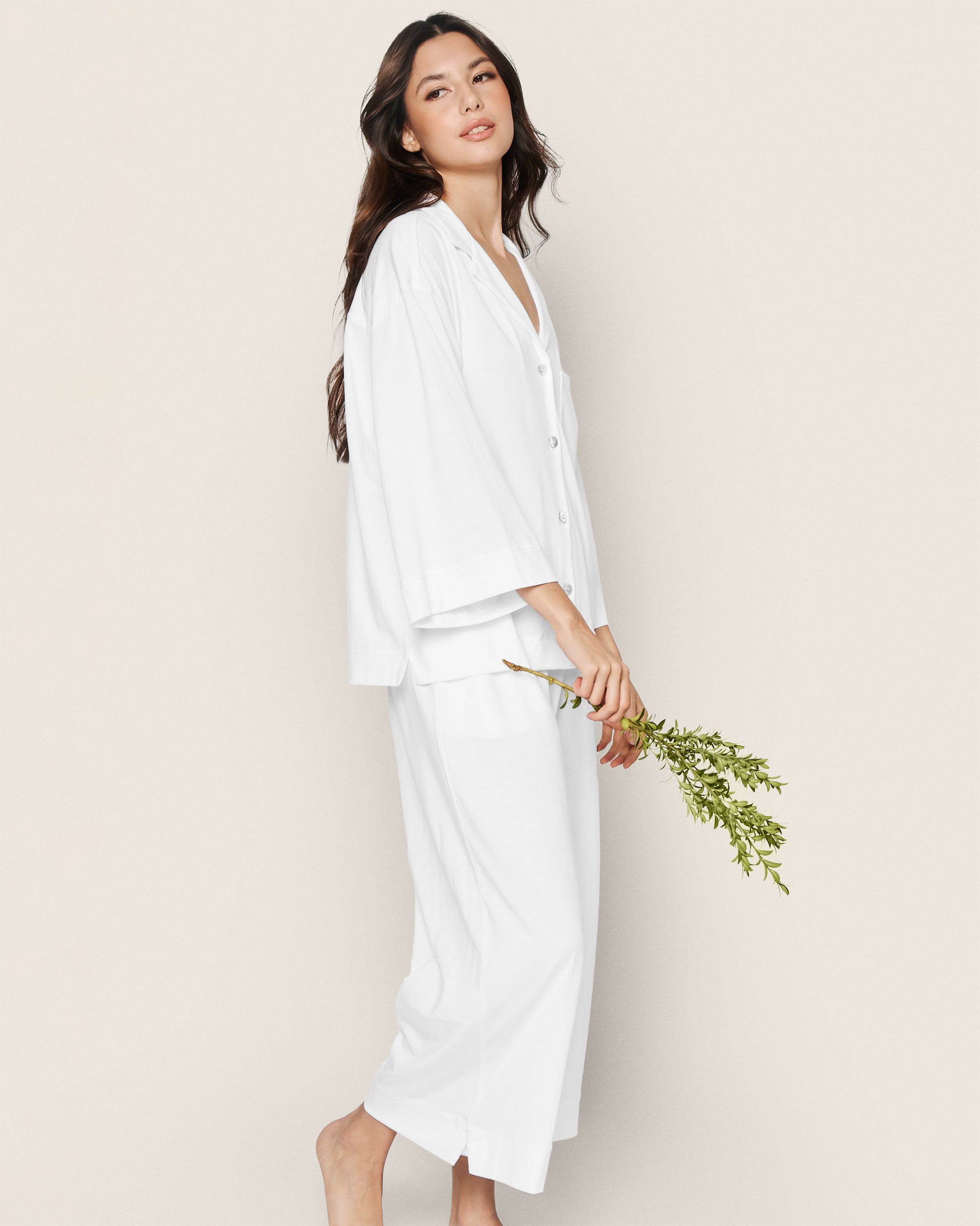 A person with long dark hair is wearing The Olivia Wide Leg Pima Pajama Set in White by Petite Plume. Crafted from soft Peruvian Pima cotton, they stand barefoot against a beige background, holding a small greenery branch and gazing slightly to the side.