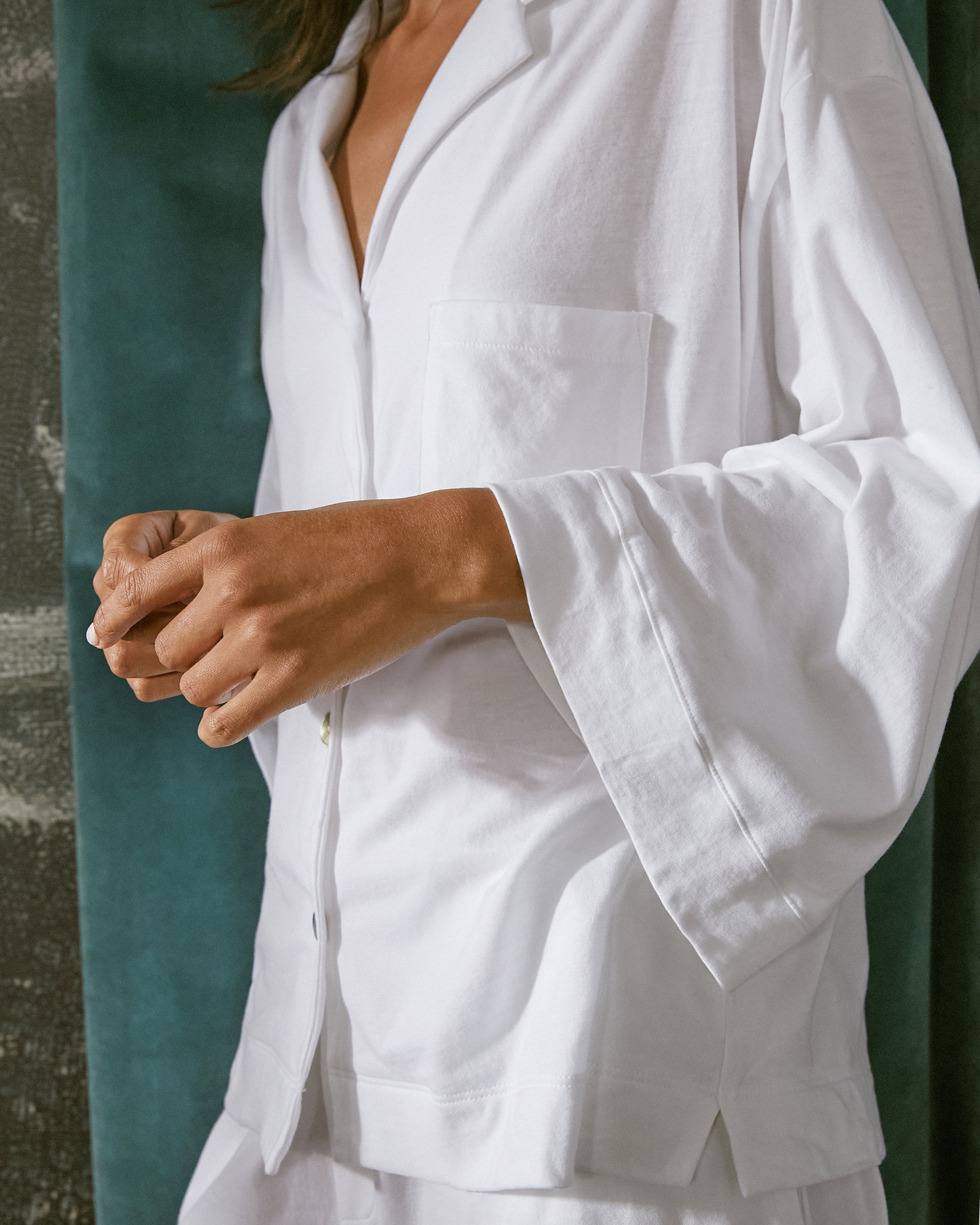 The person is wearing the white Olivia Wide Leg Pima Pajama Set by Petite Plume, featuring loose-fitting wide sleeves in Peruvian Pima cotton. Their left hand gently touches their right sleeve against a partially visible textured dark green background.