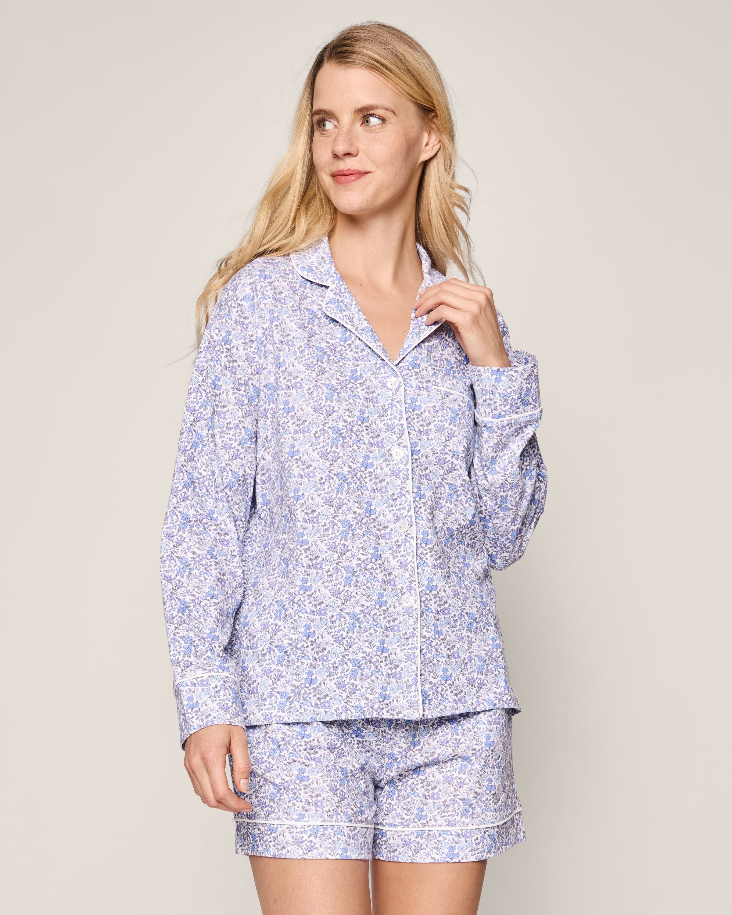 A woman with long blonde hair wears the Womens Pima Long Sleeve Short Set in Fleur dAzur by Petite Plume. The vintage-inspired blue and white floral pajama set features a long-sleeve shirt and mid-thigh shorts. She stands against a plain background, smiling slightly and looking off to the side.