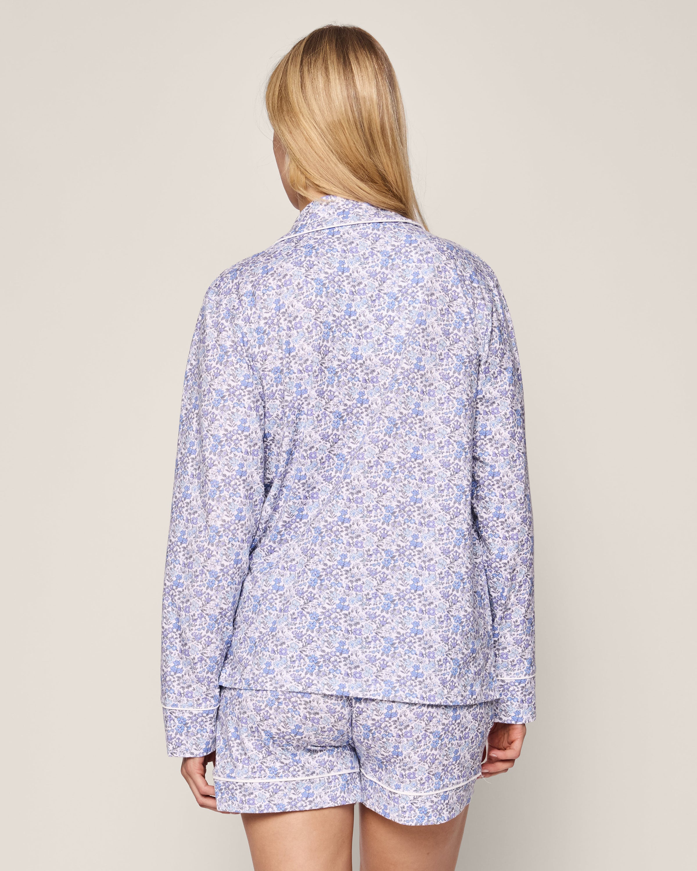 A person with long blonde hair is seen from behind in Petite Plumes Womens Pima Long Sleeve Short Set in Fleur dAzur, featuring a vintage-inspired blue floral print. The set includes a long-sleeve top and mid-thigh shorts, against a plain light background.