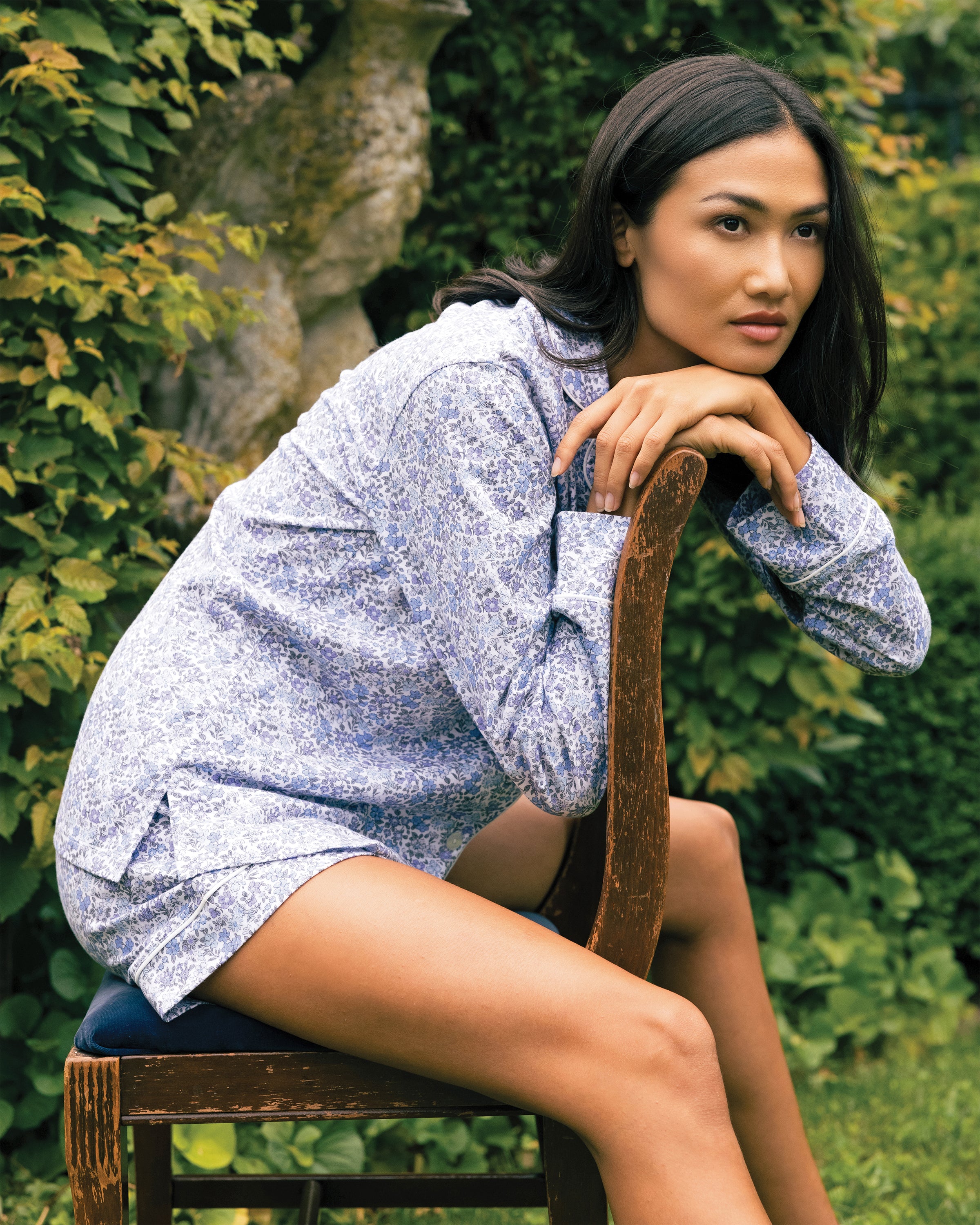 A woman in Petite Plumes Womens Pima Long Sleeve Short Set in Fleur dAzur sits backwards on a wooden chair, resting her arms and chin on its back. She gazes to her right, against a lush, green foliage backdrop.