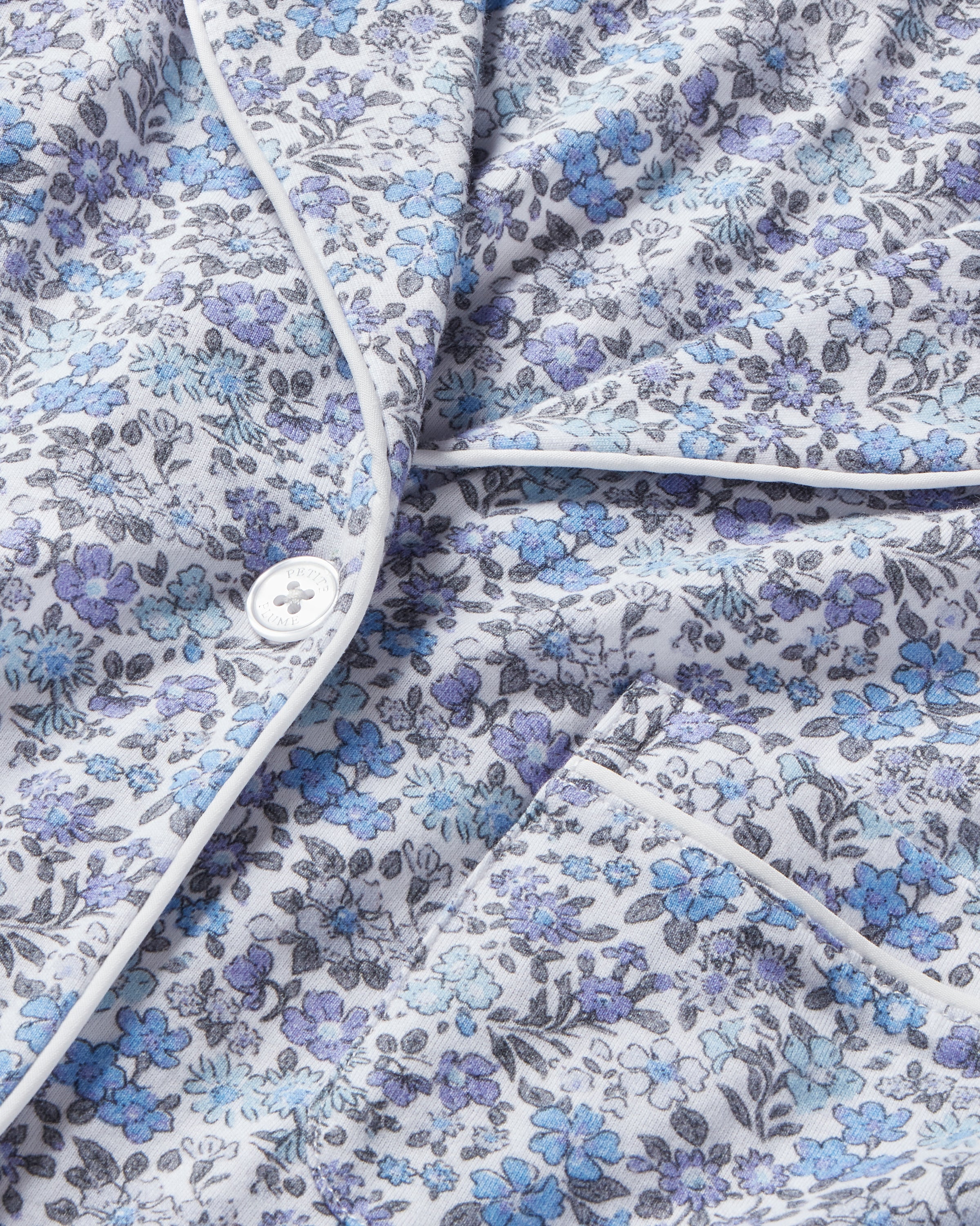 Close-up of a vintage-inspired print on Petite Plume’s Womens Pima Long Sleeve Short Set in Fleur dAzur featuring small blue and purple flowers on light fabric, adorned with white piping, buttons, and a visible pocket—perfectly reminiscent of classic PJs.