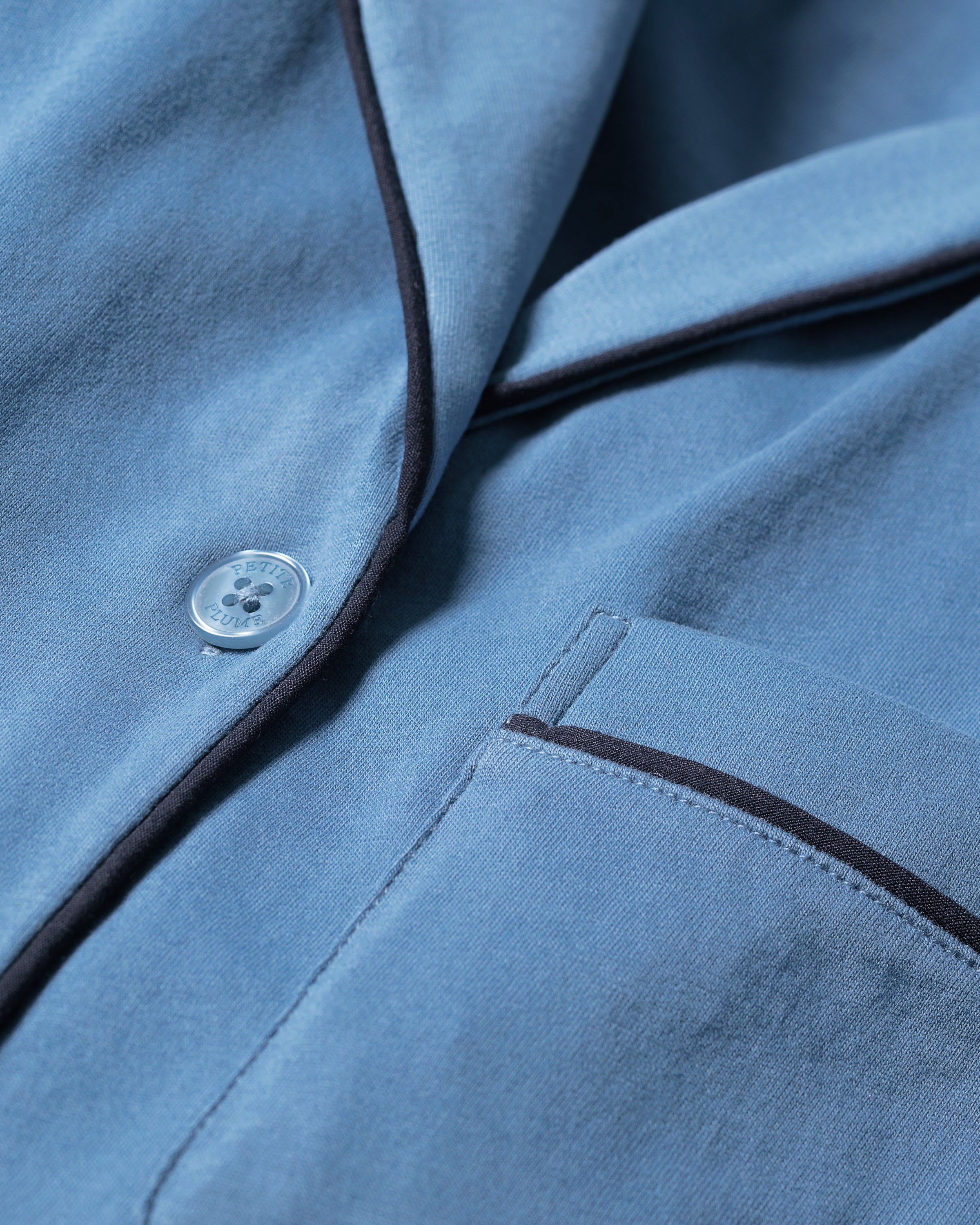 Close-up of the Womens Pima Long Sleeve Short Set in Mystique Blue by Petite Plume, showcasing serene blue luxury sleepwear with button closure, piping details, and a pocket. The Pima cotton offers a smooth texture and subtle sheen, with black trim accentuating seams and pockets.