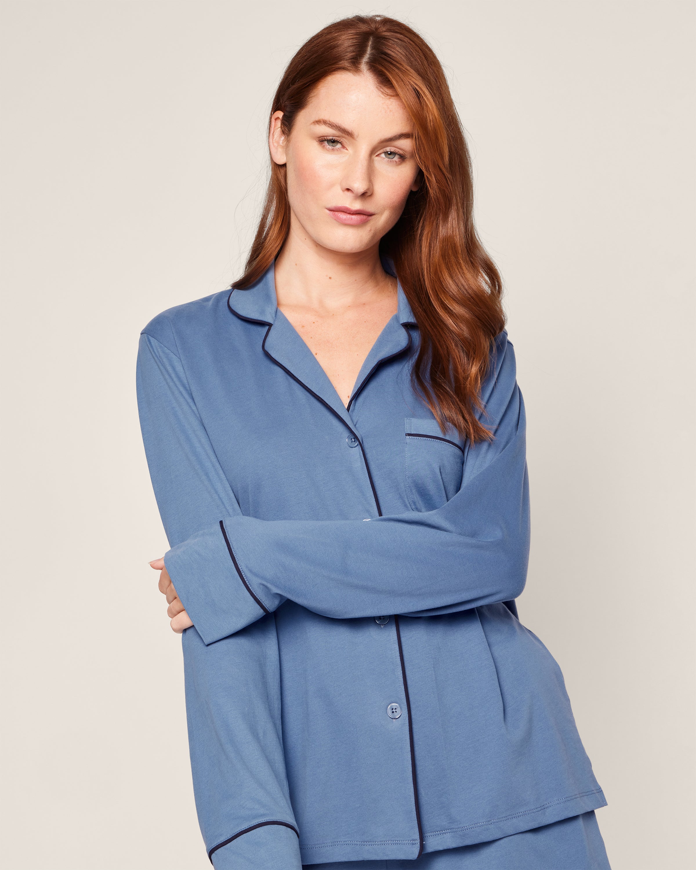 A person with long brown hair wears the Womens Pima Long Sleeve Short Set in Mystique Blue by Petite Plume, featuring a serene blue button-up pajama top with black trim. They stand against a plain background, gazing at the camera with arms confidently crossed.