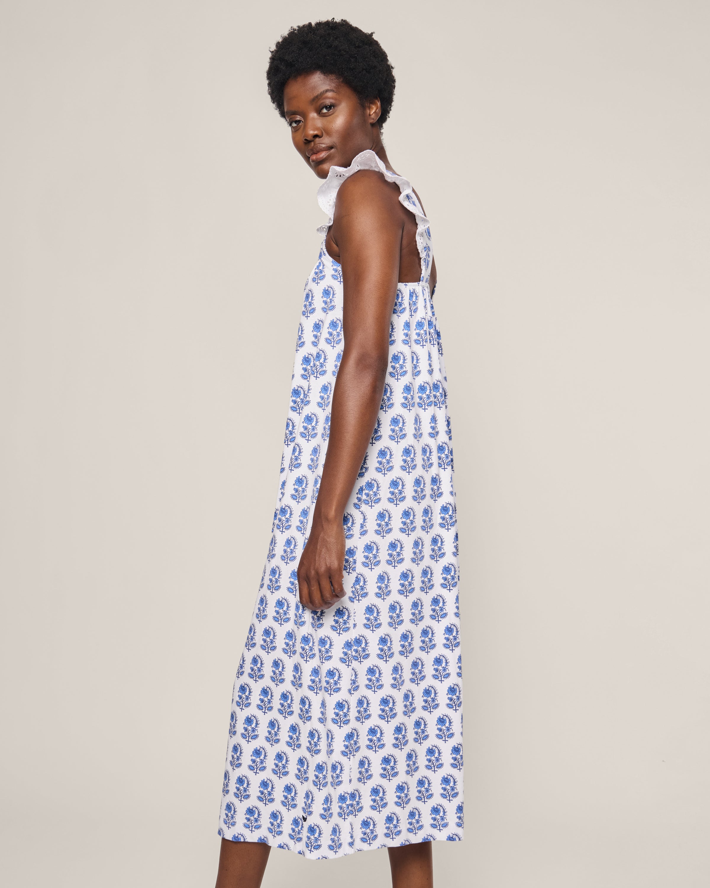 Wearing the Womens Pima Clara Nightgown in Bristol Bay by Petite Plume, a woman poses against a plain backdrop, glancing over her shoulder to highlight the luxurious blue floral pattern on the elegant white sleeveless gown.