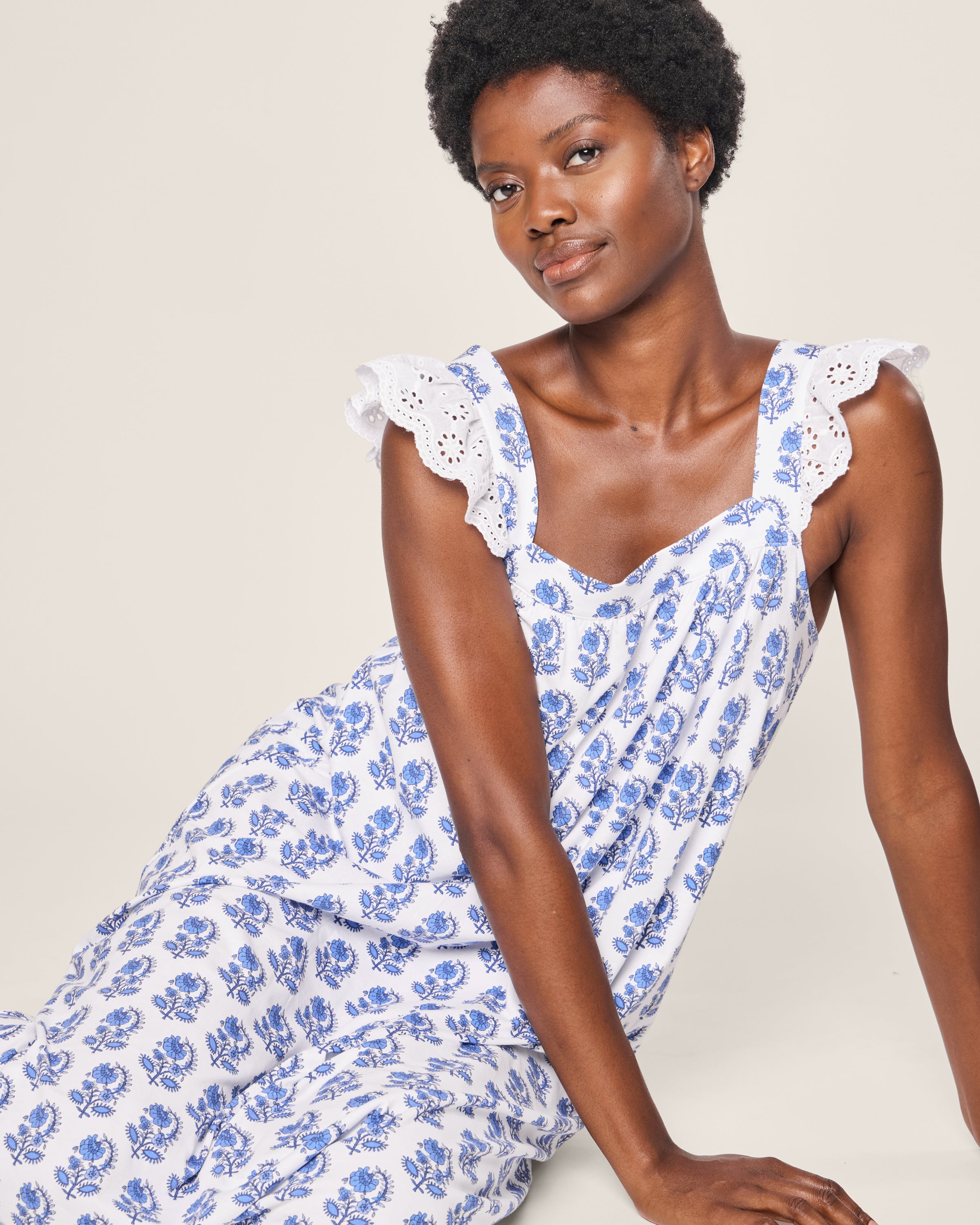 A person sits, leaning slightly, in Petite Plumes Womens Pima Clara Nightgown in Bristol Bay, featuring blue floral patterns and lace-trimmed straps. They have short curly hair and a calm expression against a plain light background.