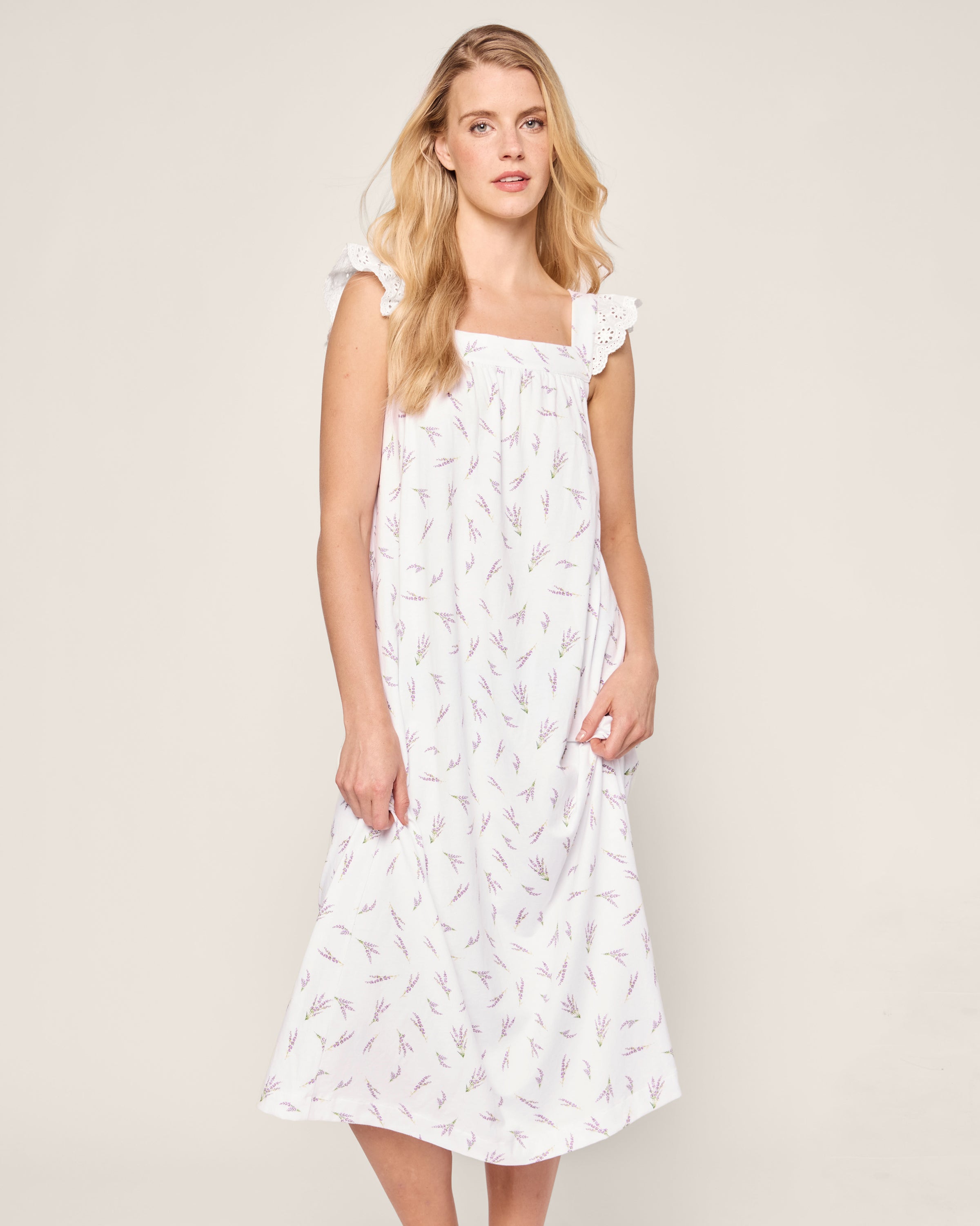 A woman with long blonde hair wears the Womens Pima Clara Nightgown in Fields of Provence by Petite Plume, featuring ruffled straps, a square neckline, and lavender floral patterns. She stands against a plain background, embodying its effortless elegance.