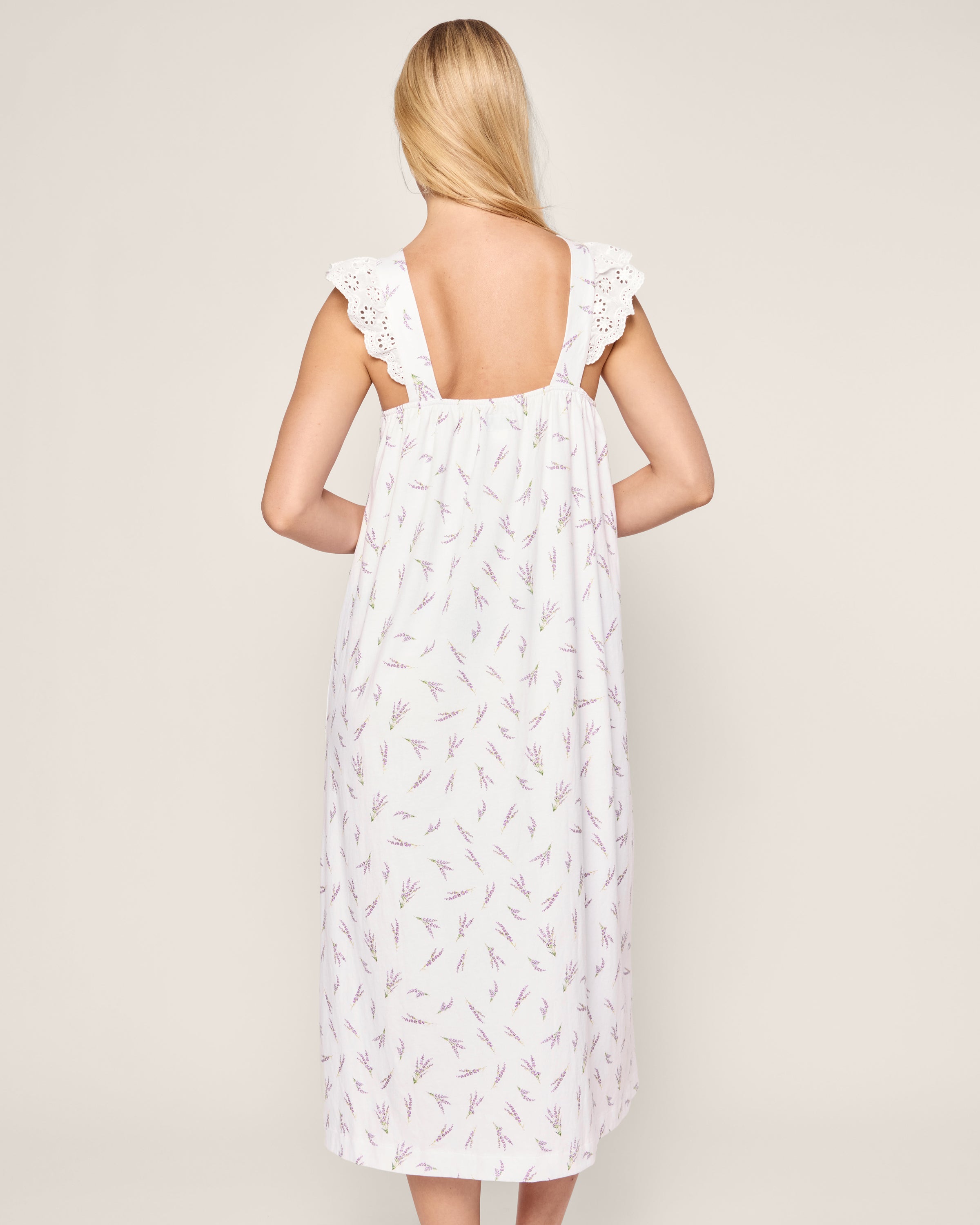 A person with long blonde hair faces away, wearing the Womens Pima Clara Nightgown in Fields of Provence by Petite Plume. This gown features a white sleeveless design with lavender flowers, lace trim, a loose fit, and ruffled shoulders against a plain light background.