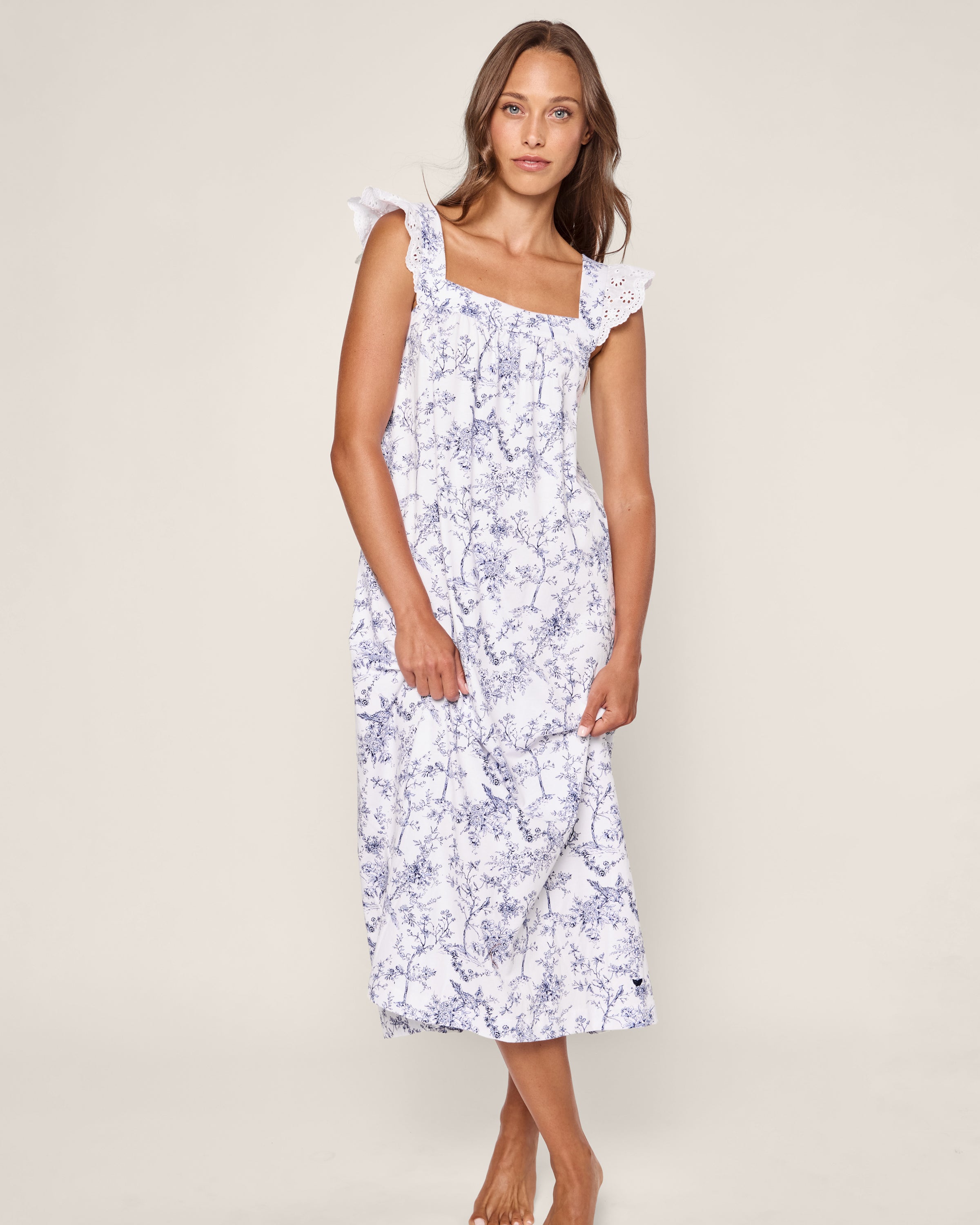 A woman in Petite Plumes Pima Clara Nightgown in Timeless Toile stands barefoot against a plain backdrop, showcasing the elegance of luxurious pajamas with its sleeveless, long white jersey knit and blue floral patterns.