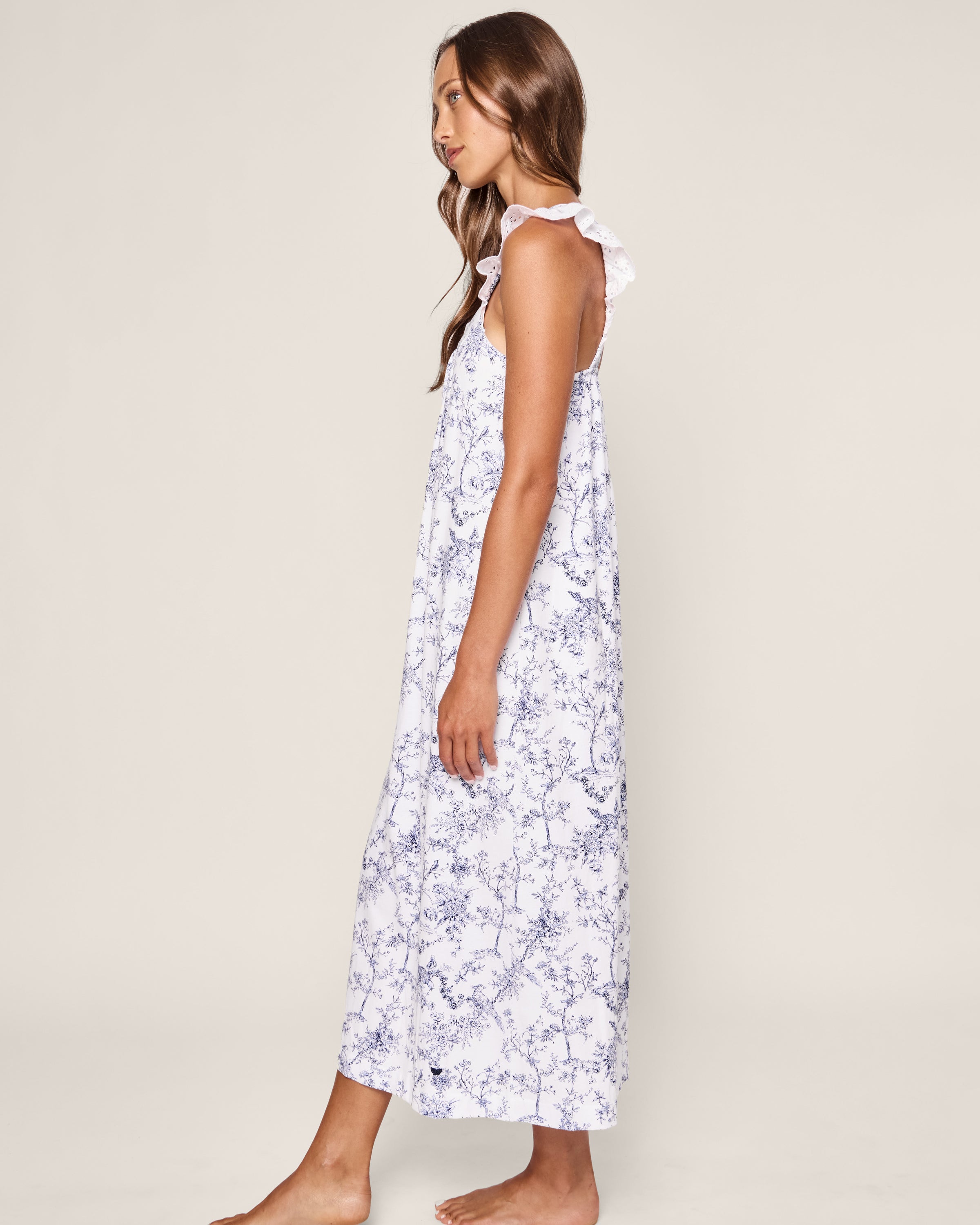 A woman with long hair stands barefoot against an off-white background, embracing her bedtime serenity. She wears Petite Plumes Womens Pima Clara Nightgown in Timeless Toile, a sleeveless ankle-length white dress with blue floral patterns and ruffled straps, facing sideways.