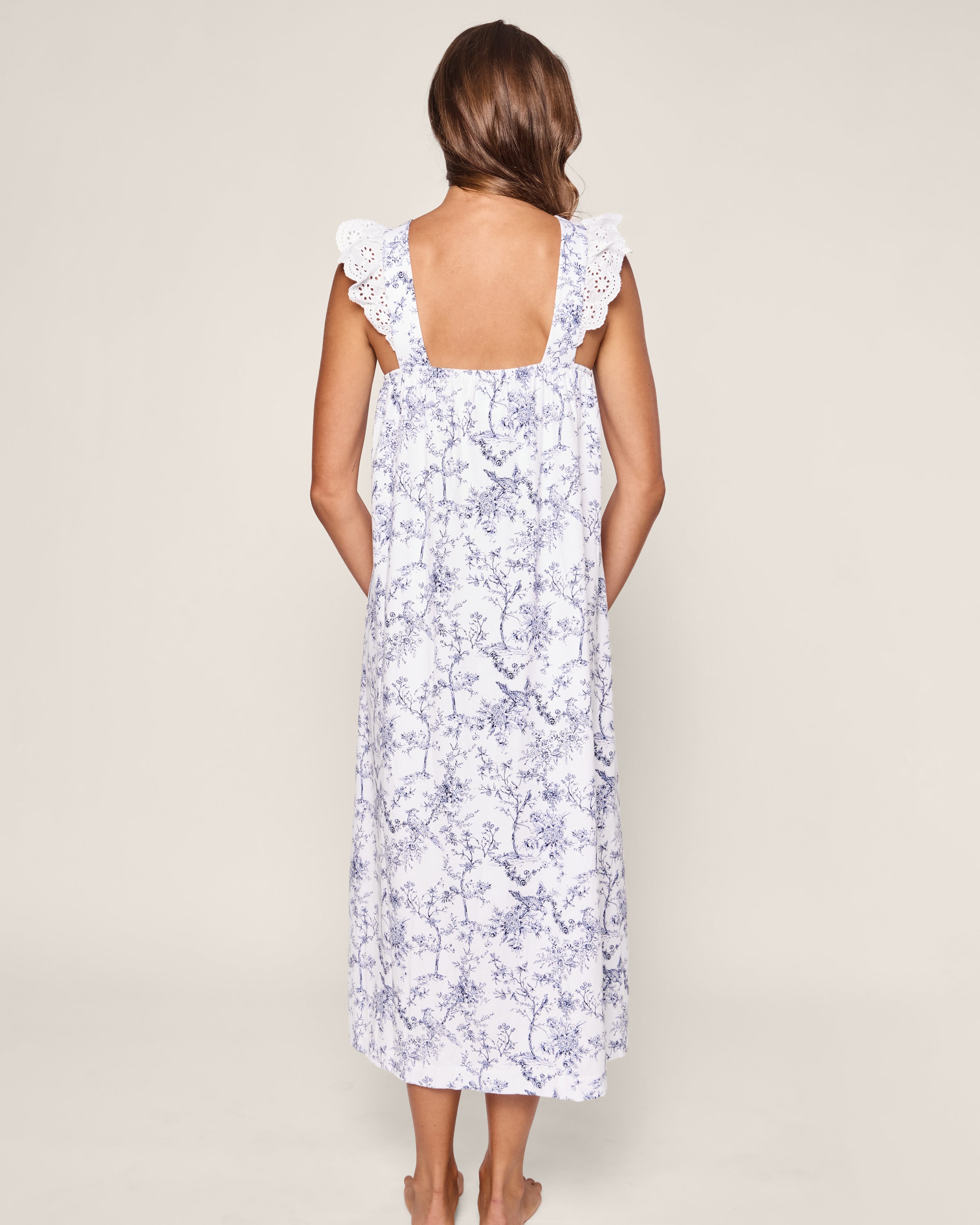 A long-haired person is depicted from the back wearing Petite Plumes Womens Pima Clara Nightgown in Timeless Toile. The sleeveless, white and blue floral dress with lace shoulder details drapes elegantly to ankle-length against a light gray background, evoking a serene bedtime ritual.