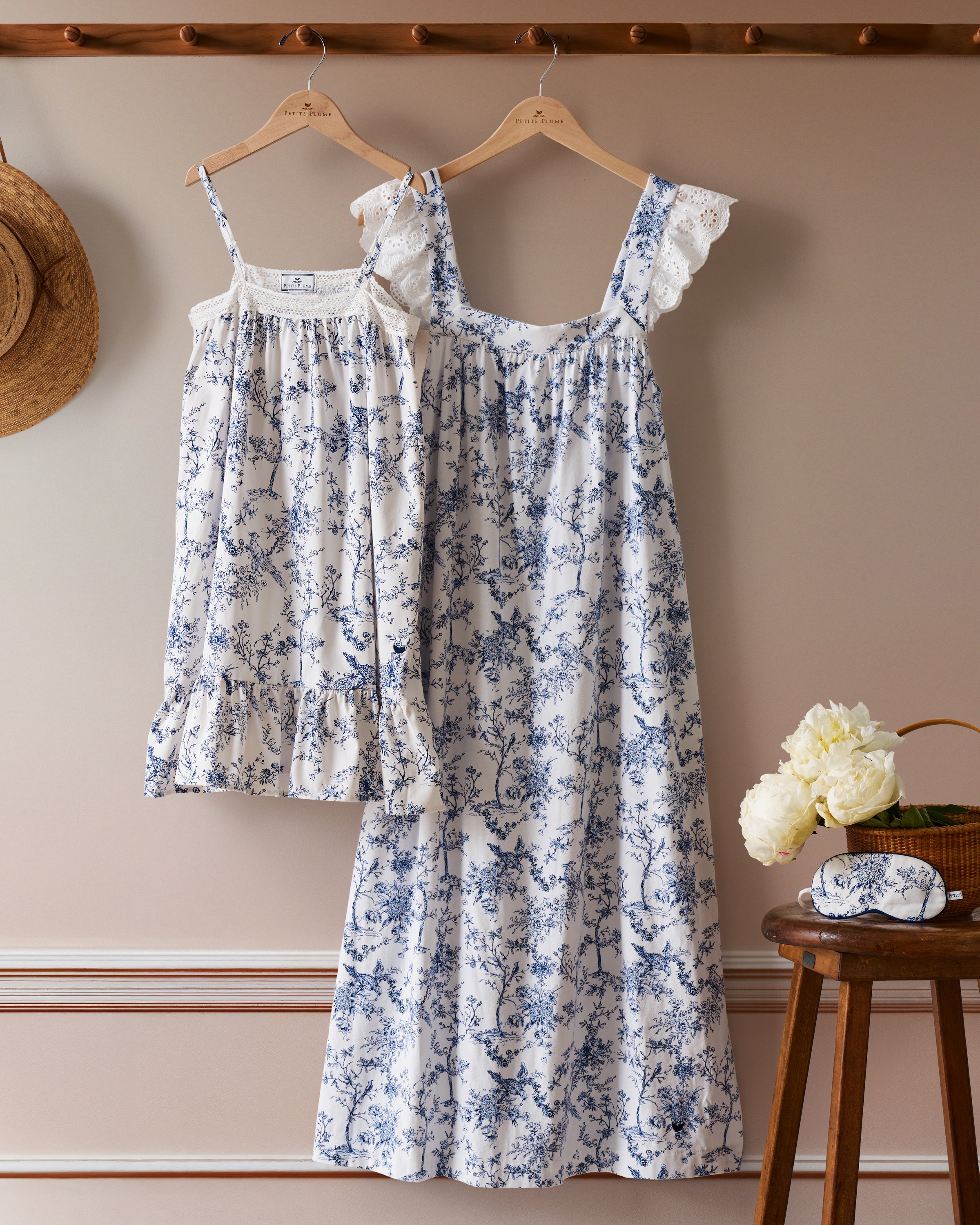 Two Womens Pima Clara Nightgowns in Timeless Toile by Petite Plume hang on wooden hangers against a beige wall. A straw hat graces the left, while a stool with white flowers and a sleep mask completes your bedtime ritual on the right.