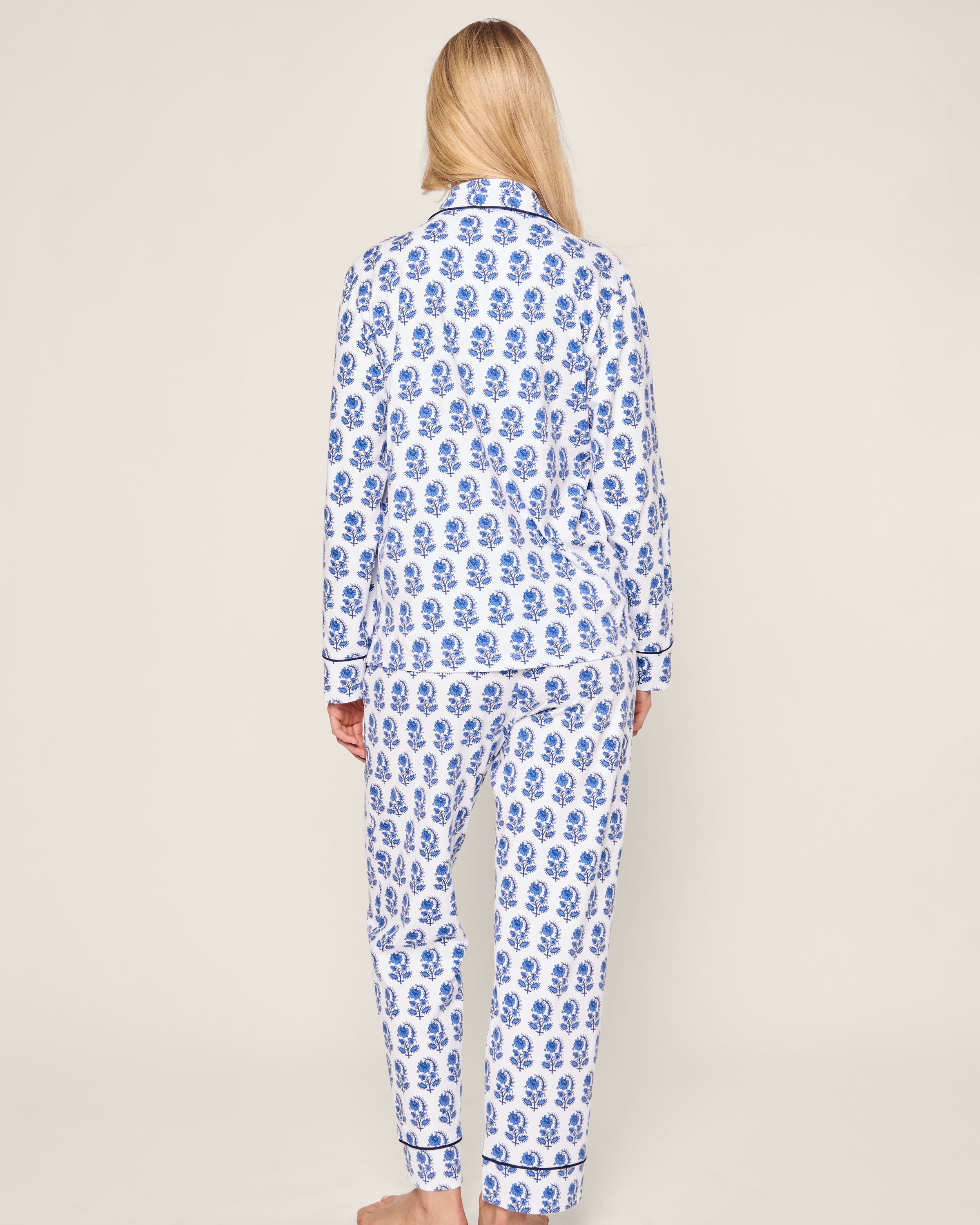 A barefoot person with long blonde hair is facing away, wearing the Petite Plume Womens Pima Pajama Set in Bristol Bay, made of luxury Peruvian Pima cotton and featuring blue and white graphic florals on a light background.