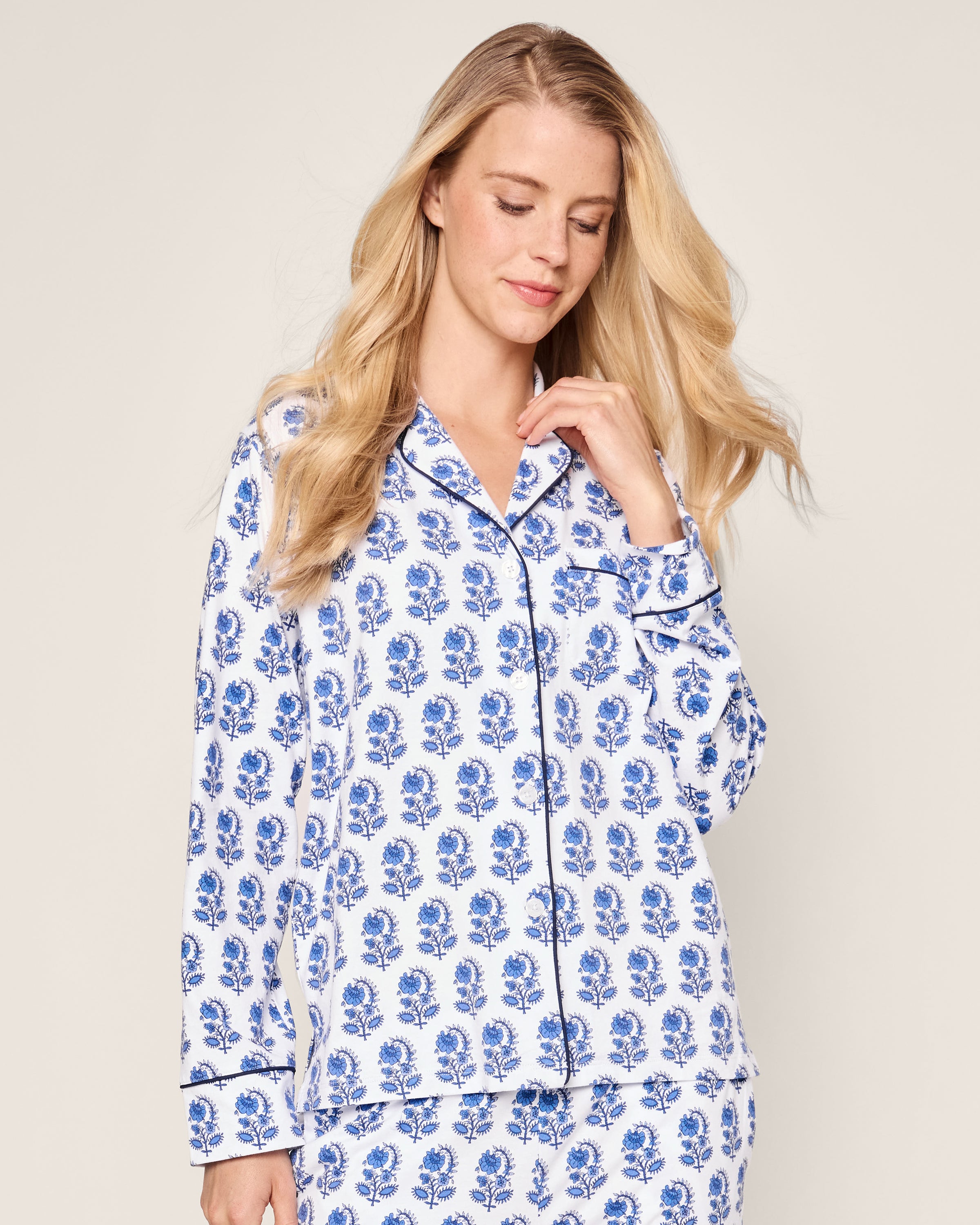 A person wearing Petite Plumes Womens Pima Pajama Set in Bristol Bay, featuring blue and white graphic florals made from Peruvian Pima cotton, with long sleeves and a collar. They have long, wavy blonde hair and a slight smile against a plain background.