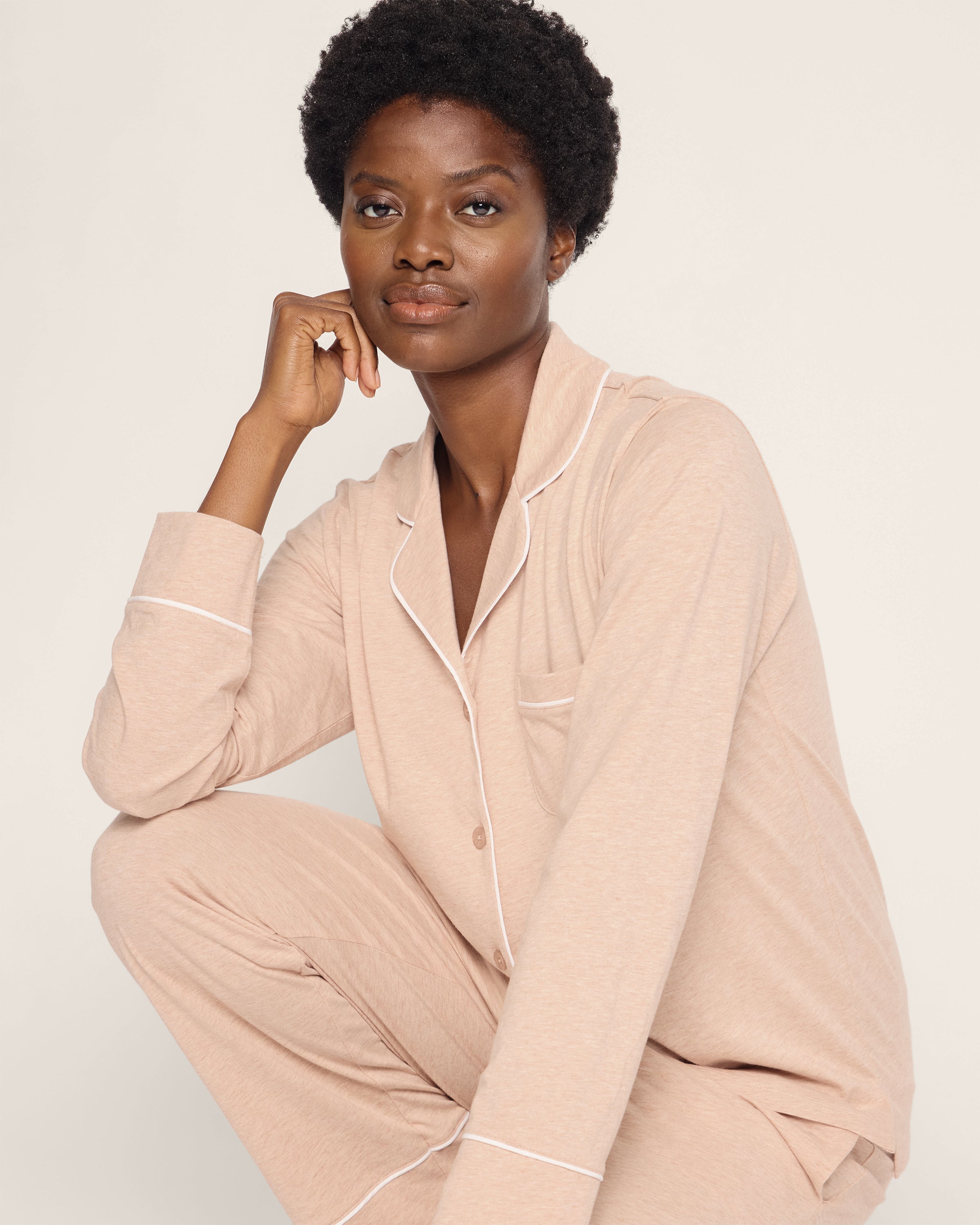 A person with short curly hair wears Petite Plumes Womens Pima Pajama Set in Camel while sitting with one knee up, chin on hand, gazing at the camera. The off-white background enhances the elegance of their luxury sleepwear.