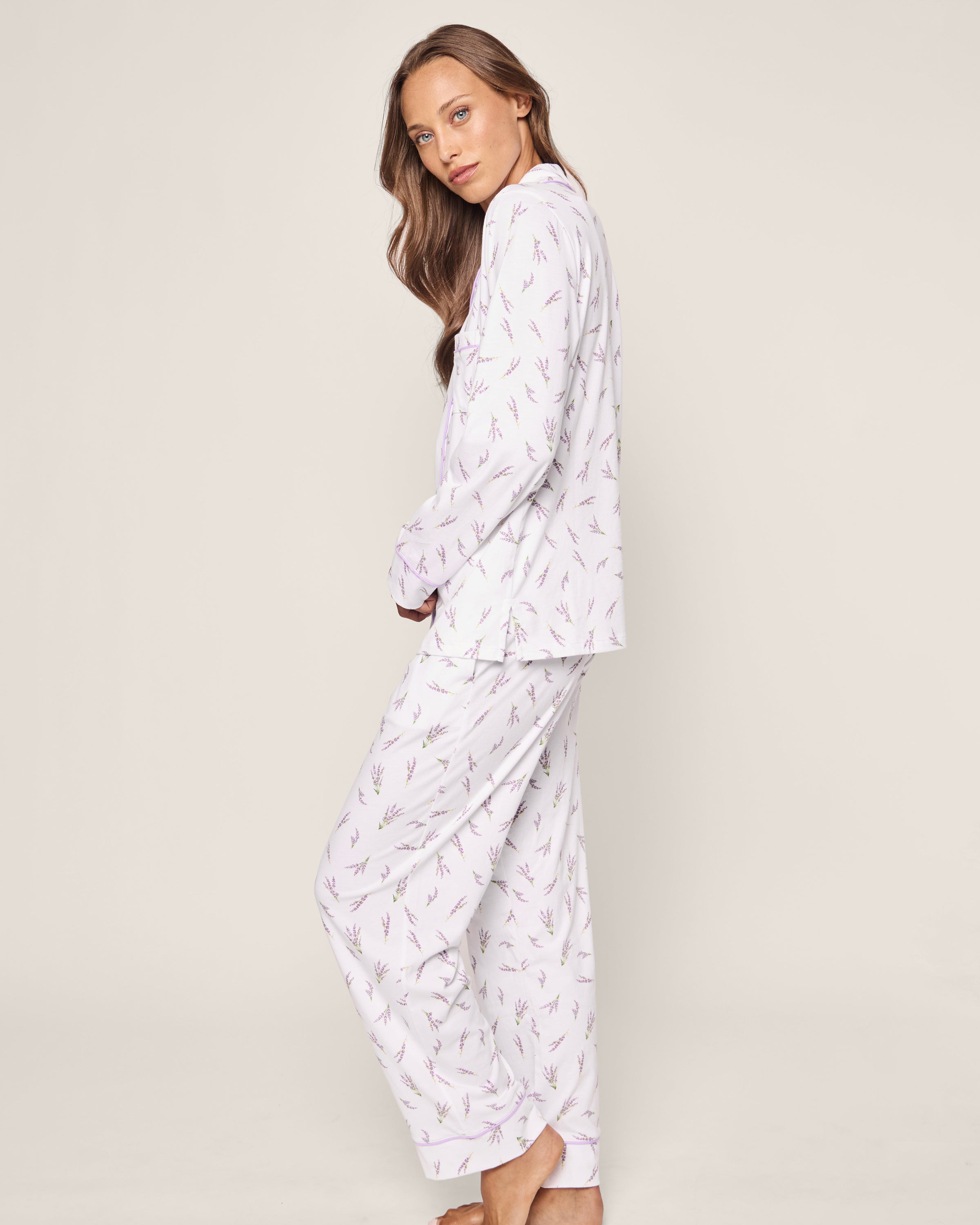 A woman with long brown hair wears the Womens Pima Pajama Set in Fields of Provence by Petite Plume. The luxurious sleepwear, elegantly patterned with lavender flowers, captures a timeless silhouette as she stands barefoot on a plain background, her eyes meeting the camera.