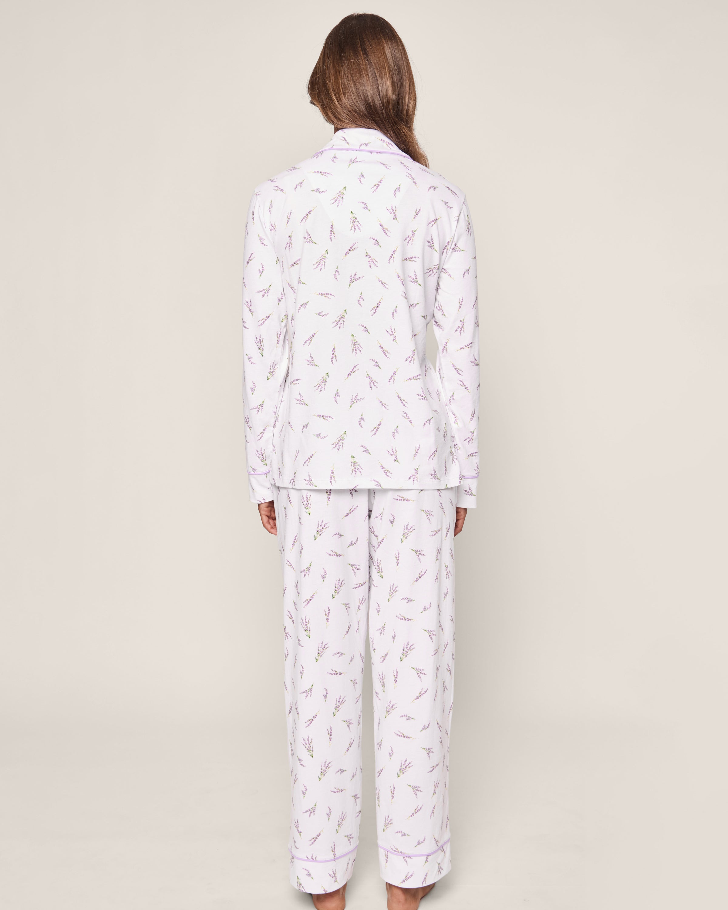 A person with long brown hair faces away, wearing Petite Plumes Womens Pima Pajama Set in Fields of Provence. The white pajama set is adorned with lavender flowers in a classic design, complemented by a plain, light-colored background that adds elegance to the luxury sleepwear.