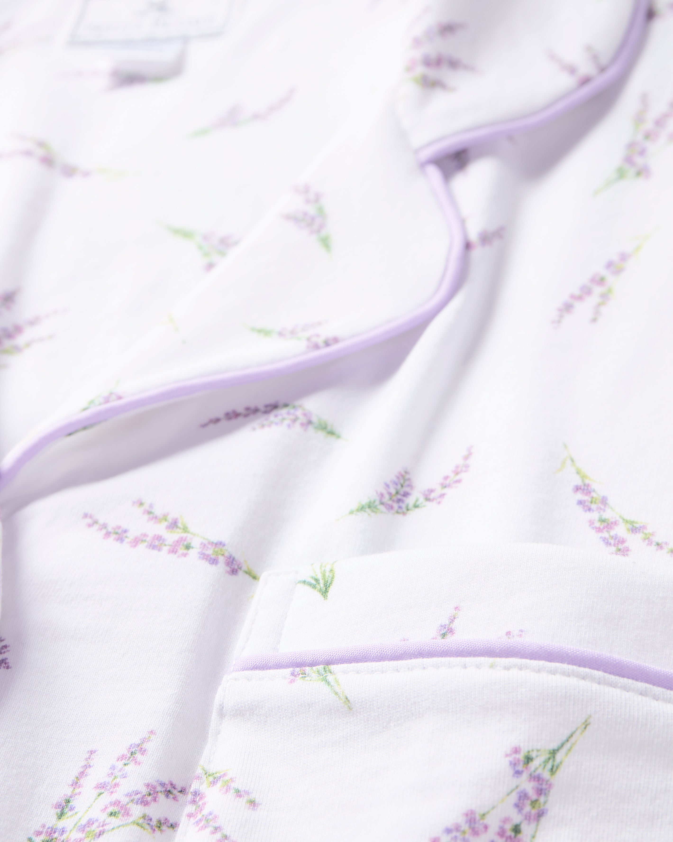 A close-up of the Womens Pima Pajama Set in Fields of Provence by Petite Plume reveals a white fabric with light purple piping and a lavender flower pattern, featuring a pocket and lapel that suggest the classic pajama silhouette.