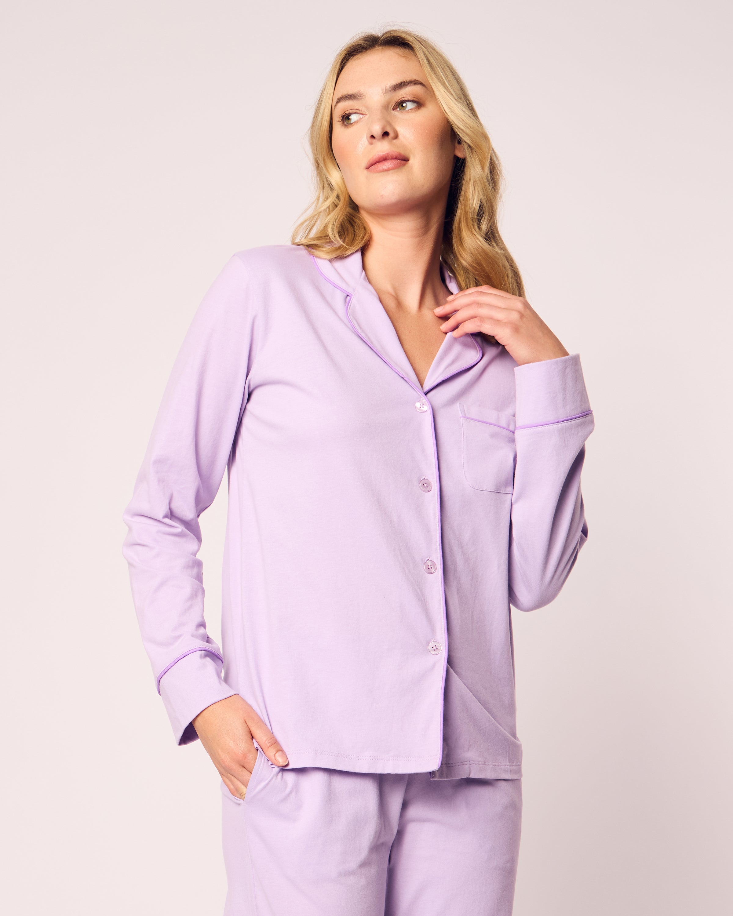 A person with long hair is wearing the Petite Plume Womens Pima Pajama Set in Lavender, featuring a luxurious button-up top with a pocket and matching pants. Made from Peruvian Pima cotton, they stand with one hand in a pocket and the other touching their hair, gazing slightly to the side.