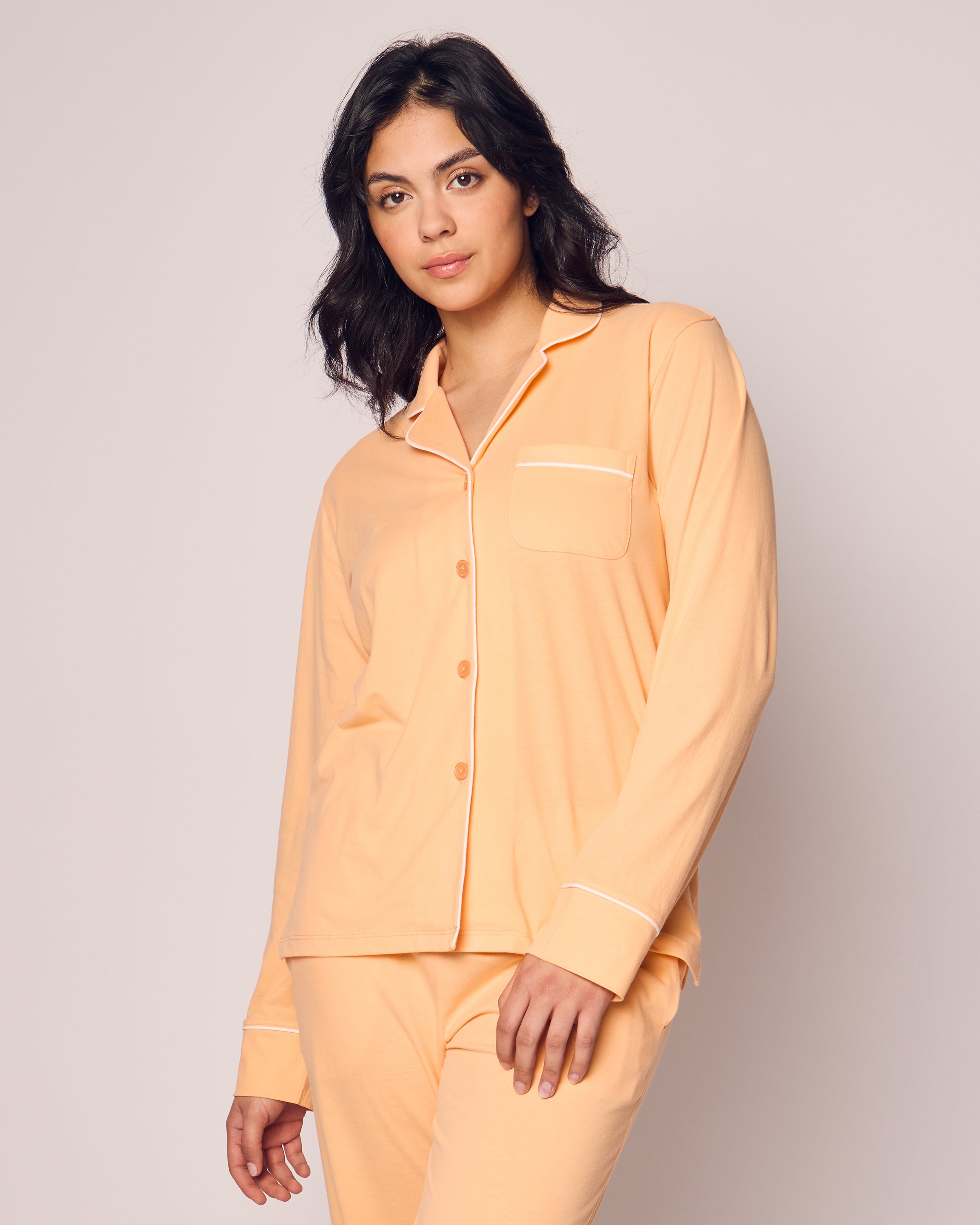 A woman models the Petite Plume Womens Pima Pajama Set in Peach against a plain background. Her medium-length dark hair enhances her relaxed expression, while the Peruvian Pima cotton top’s front pocket and buttons add to its luxurious appeal.