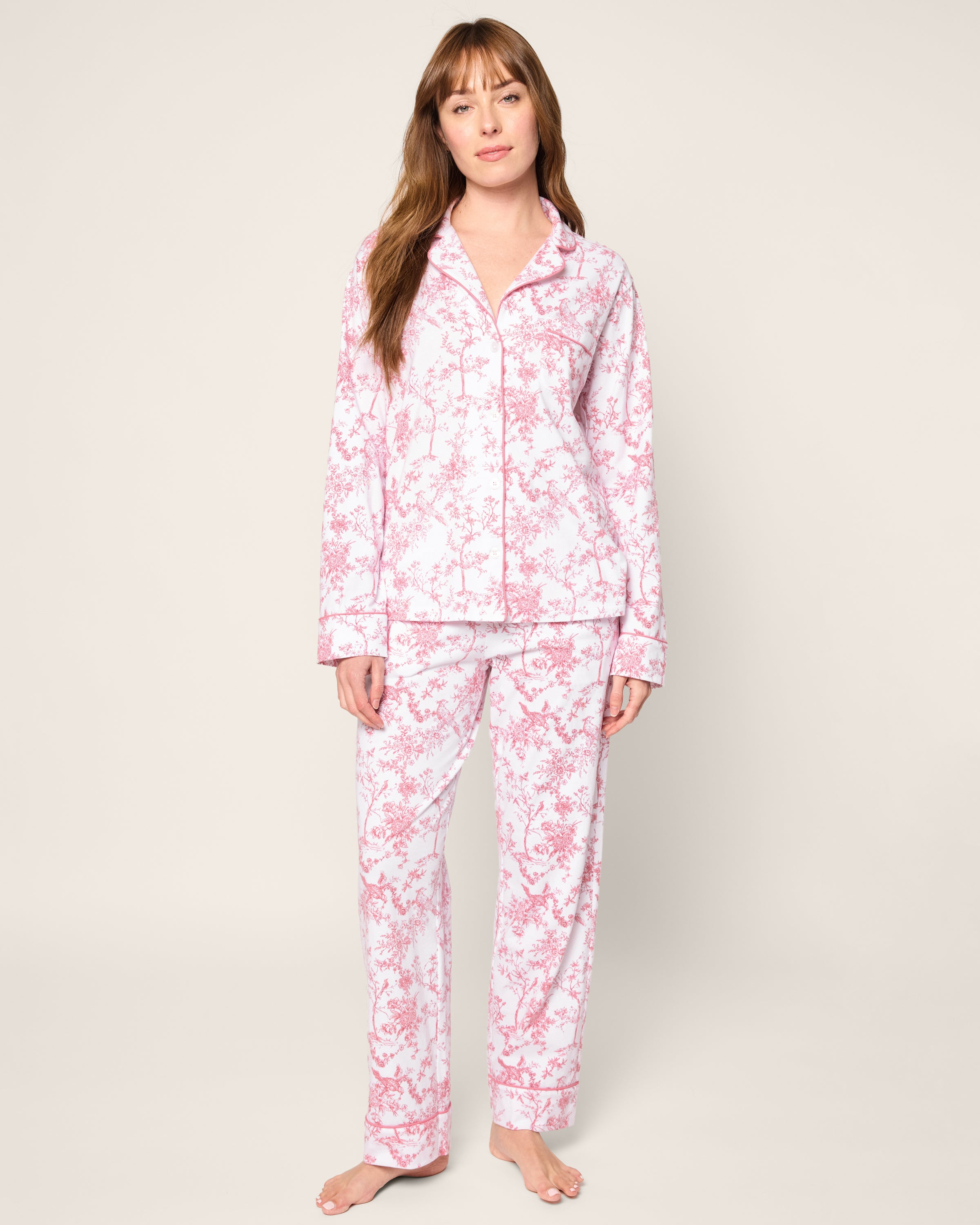 Against a light backdrop, a person stands barefoot in Petite Plumes Womens Pima Pajama in Pink Timeless Toile, featuring a pink floral design on white jersey knit. The pajama top has long sleeves and a collar, while the pants are full-length, creating an inviting bedtime scene.