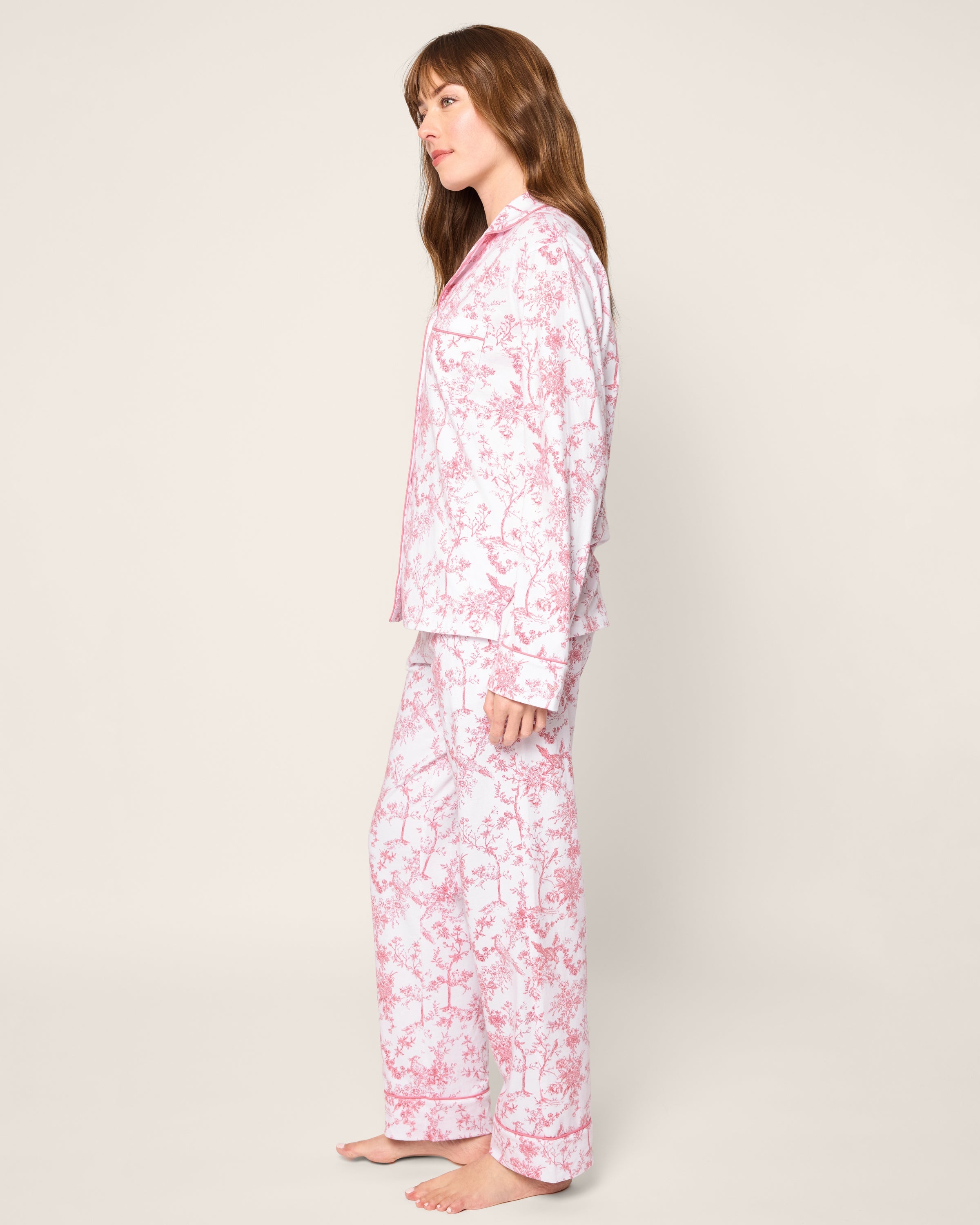 A person with long hair stands sideways in a luxurious Womens Pima Pajama in Pink Timeless Toile by Petite Plume, set against a plain, light backdrop that evokes a serene bedtime ritual.