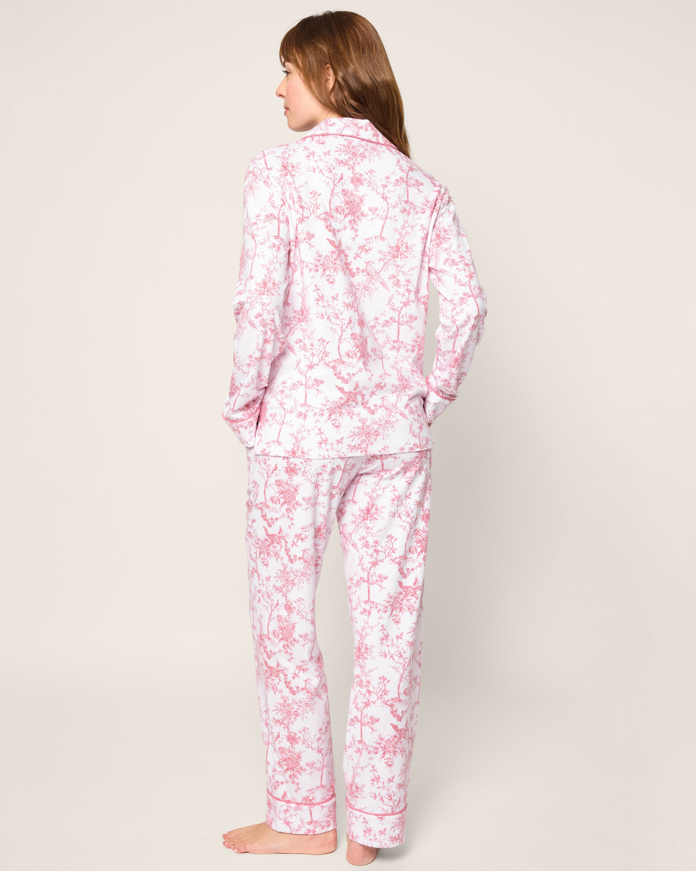 A person with long hair, barefoot and hands in pockets, is wearing Petite Plumes Womens Pima Pajama in Pink Timeless Toile. They stand with their back to the camera against a plain background.