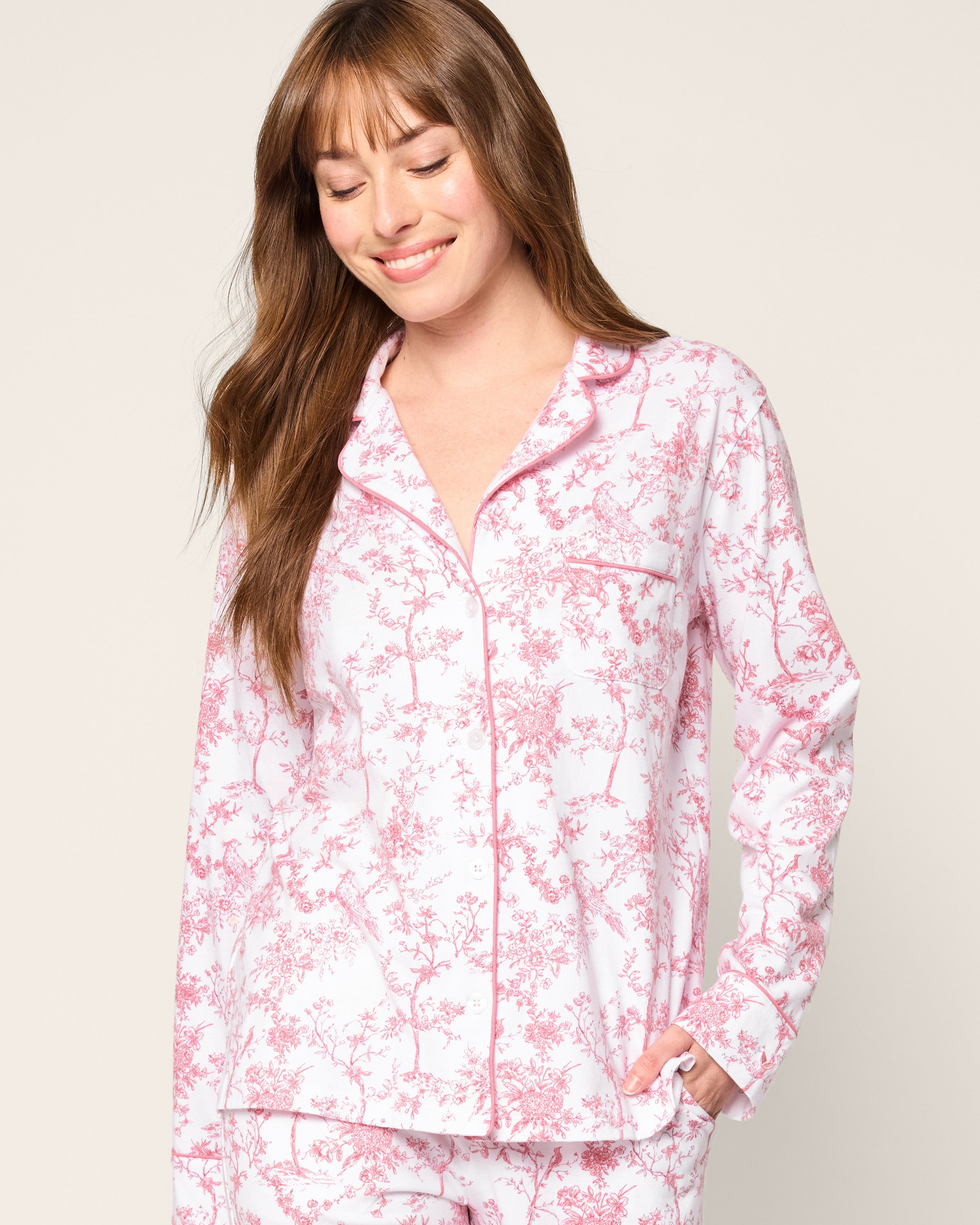 A woman with long brown hair, wearing a Petite Plume Womens Pima Pajama in Pink Timeless Toile, smiles with eyes closed and her right hand in her pocket against a plain light background.