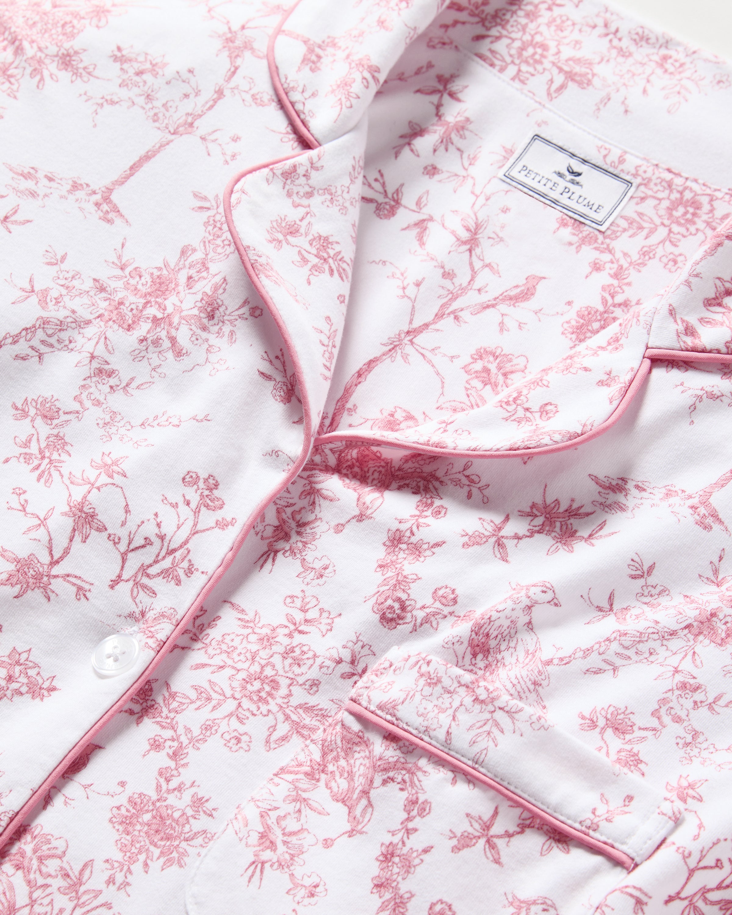 The Womens Pima Pajama in Pink Timeless Toile by Petite Plume boasts a white top adorned with pink botanical patterns, featuring pink piping on the collar, button placket, and pocket edges. Its part of the Pima Collection with a fabric label near the collar.