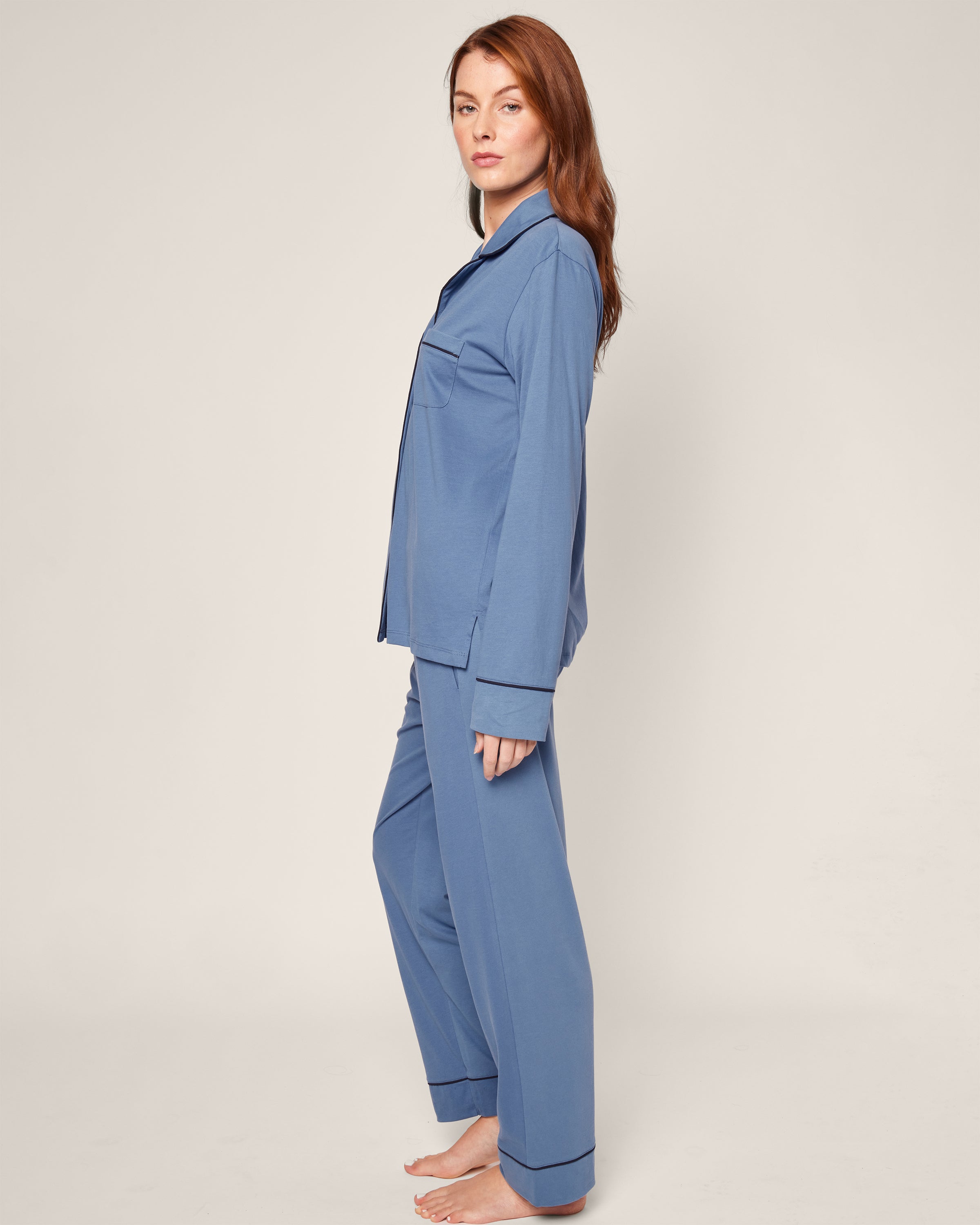 A long red-haired person stands barefoot in profile, modeling the Womens Pima Pajama Set in Mystique Blue by Petite Plume. The set features long sleeves and pants made from soft Peruvian Pima cotton, highlighted against a plain off-white background.