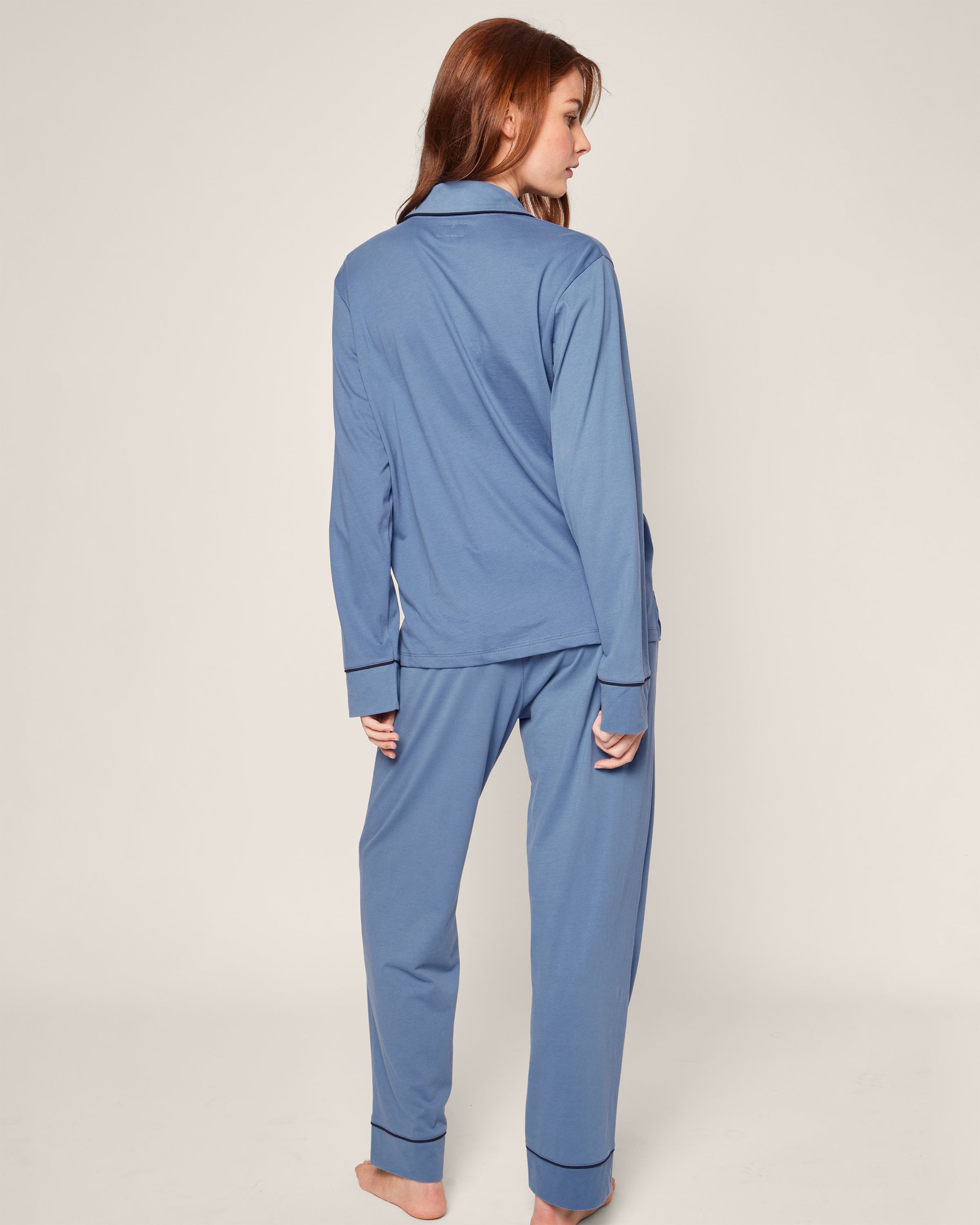 A woman with long brown hair stands barefoot, facing away, in a Petite Plume Womens Pima Pajama Set in Mystique Blue. The luxury sleepwear includes a long-sleeved top and loose-fitting pants with dark piping. She is set against a plain, light-colored background.