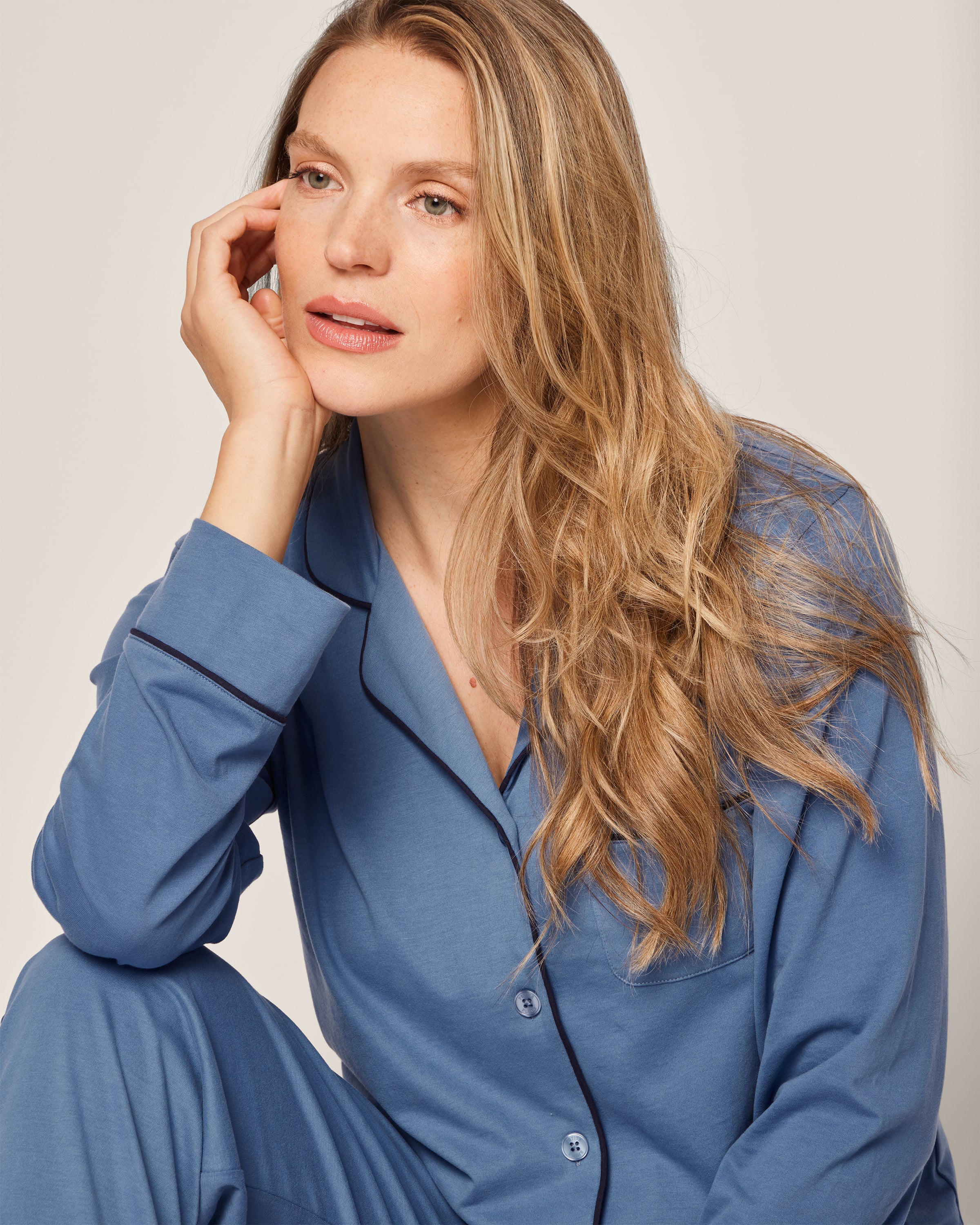 A person with long wavy hair wearing the Womens Pima Pajama Set in Mystique Blue by Petite Plume sits resting their cheek on one hand, appearing thoughtful and gazing slightly upward against a plain, light-colored background.