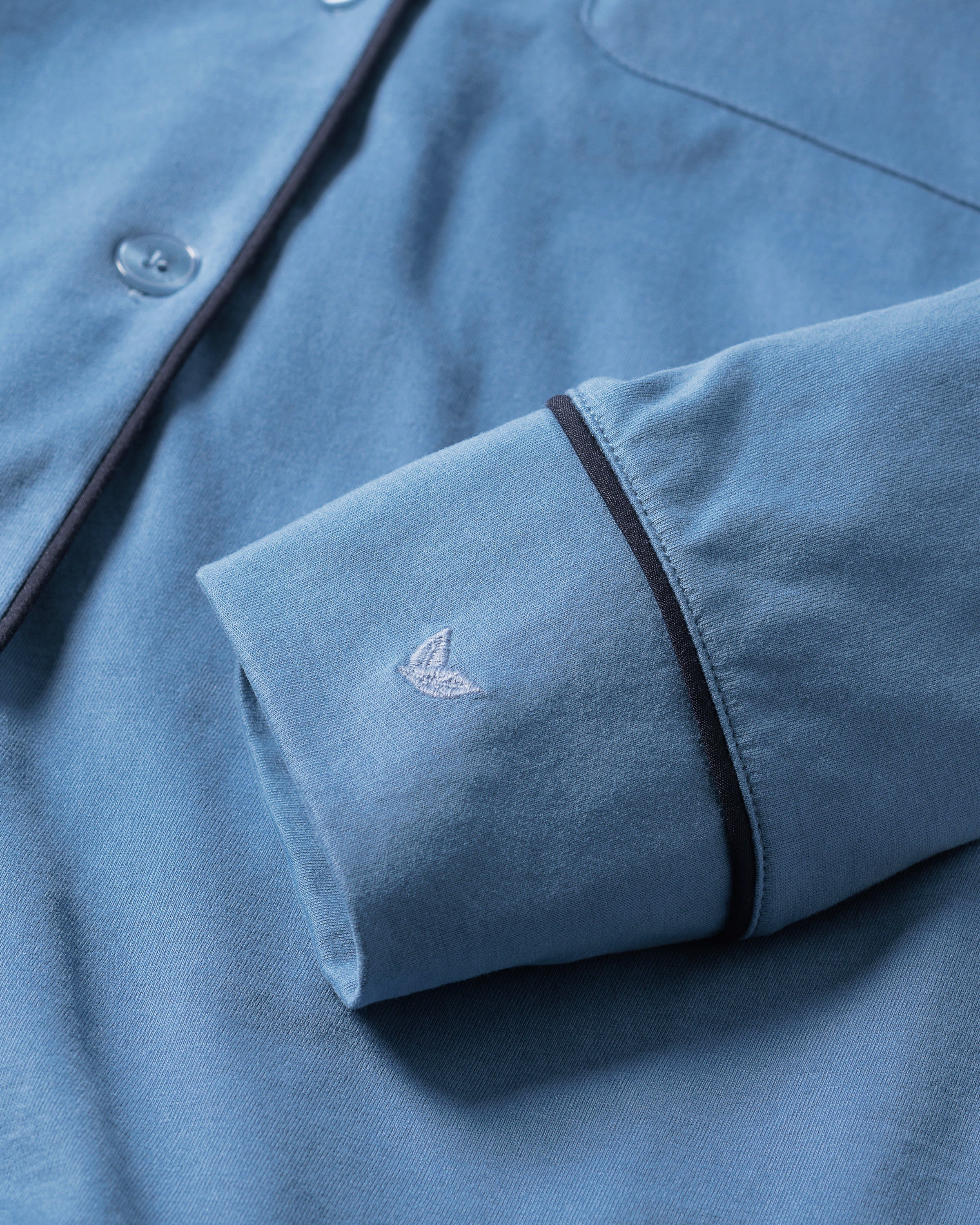 Close-up of the Womens Pima Pajama Set in Mystique Blue by Petite Plume, crafted from luxurious Peruvian Pima cotton. The sleeve has a small embroidered logo near the cuff, with black piping and blue buttons, exuding elegance. The fabric appears soft and well-tailored.