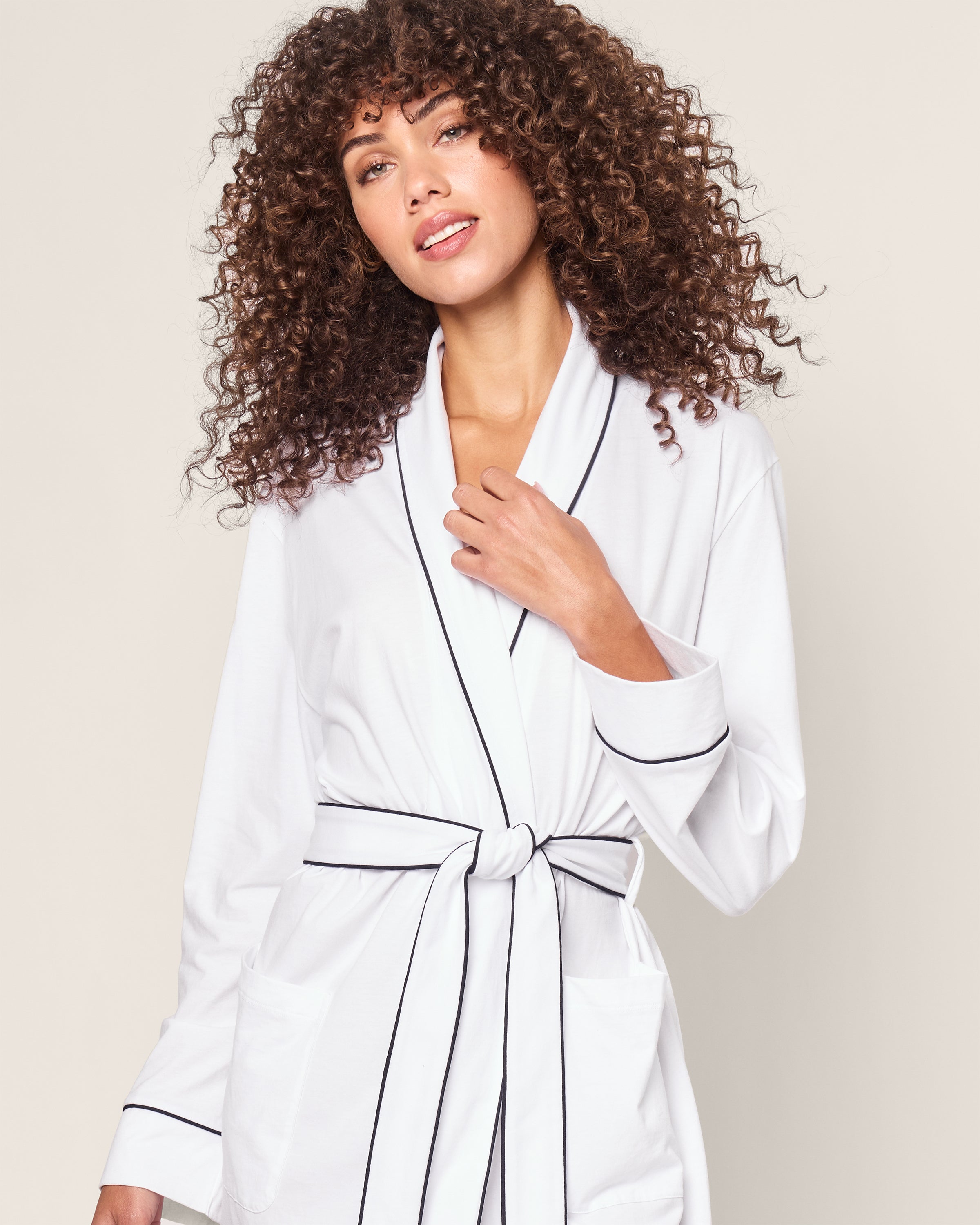 A person with curly hair models a Petite Plume Womens Pima Robe in White with Navy Piping, tied at the waist, against a neutral background. They have a calm expression.