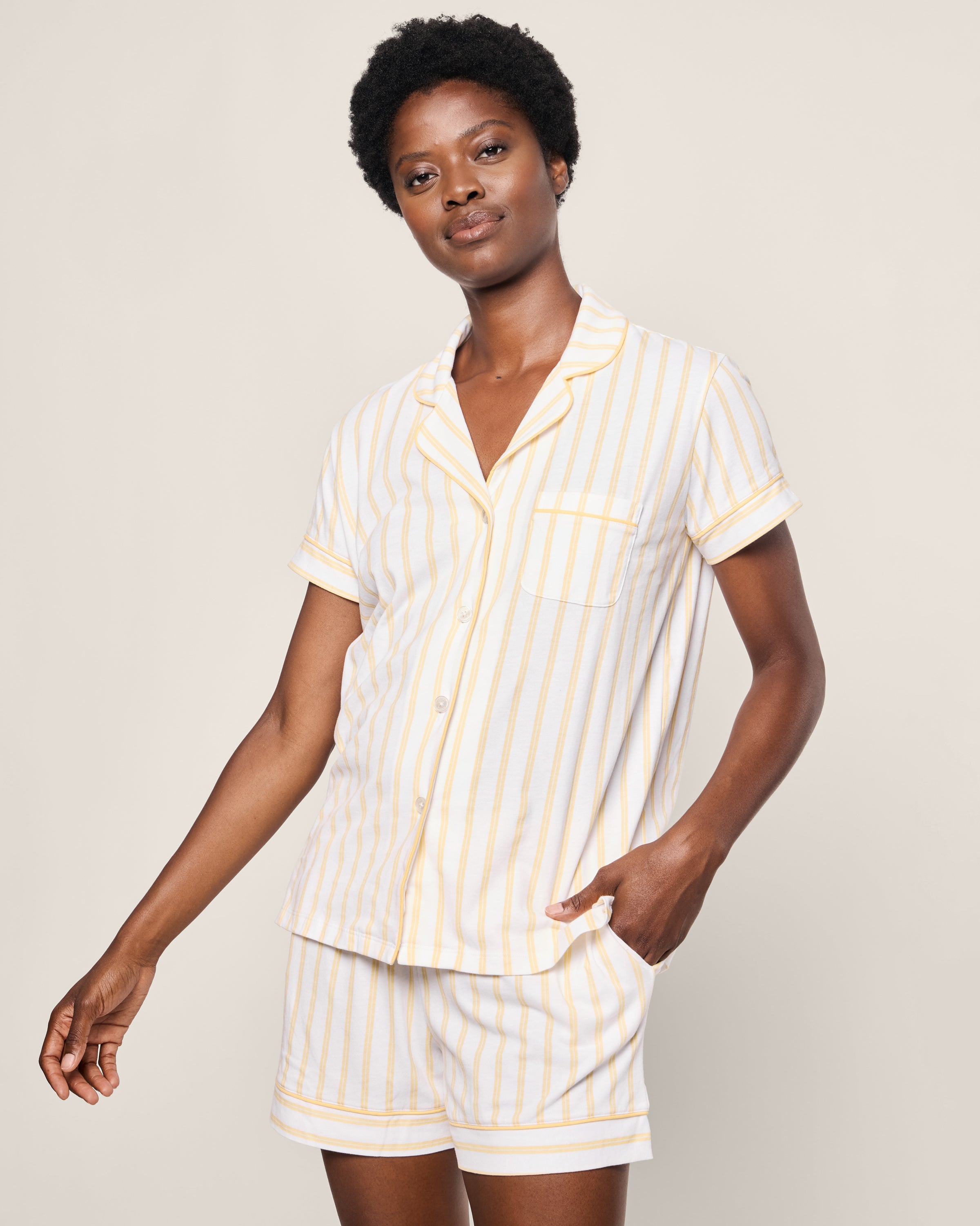 A person models the Womens Pima Short Sleeve Short Set in Sunny Stripe by Petite Plume, featuring a luxurious Peruvian Pima cotton button-up shirt and shorts with white and yellow stripes. They stand against a plain background with one hand in a pocket.