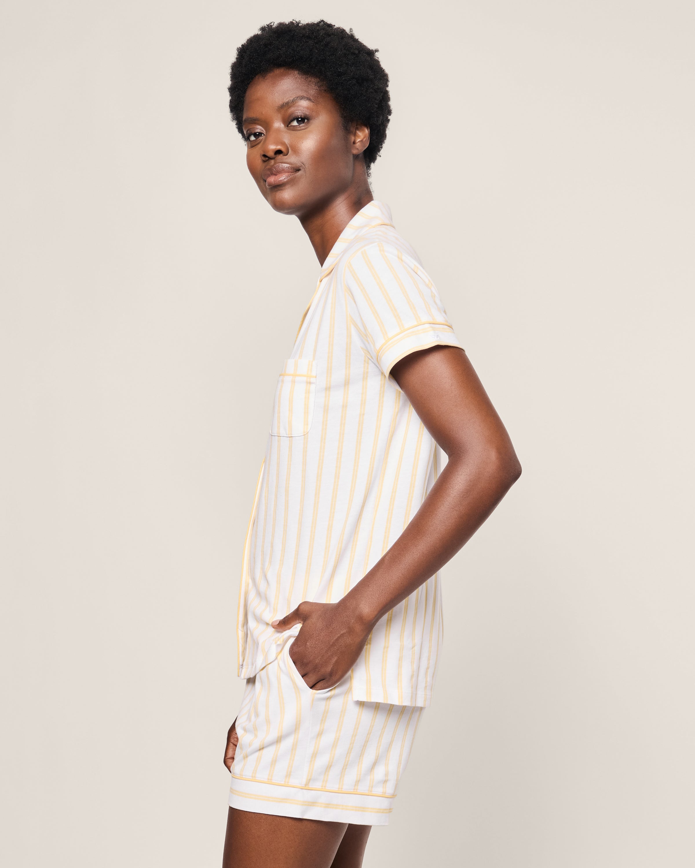 A woman wearing Petite Plumes Womens Pima Short Sleeve Short Set in Sunny Stripe stands sideways with hands in pockets, against a plain off-white background.