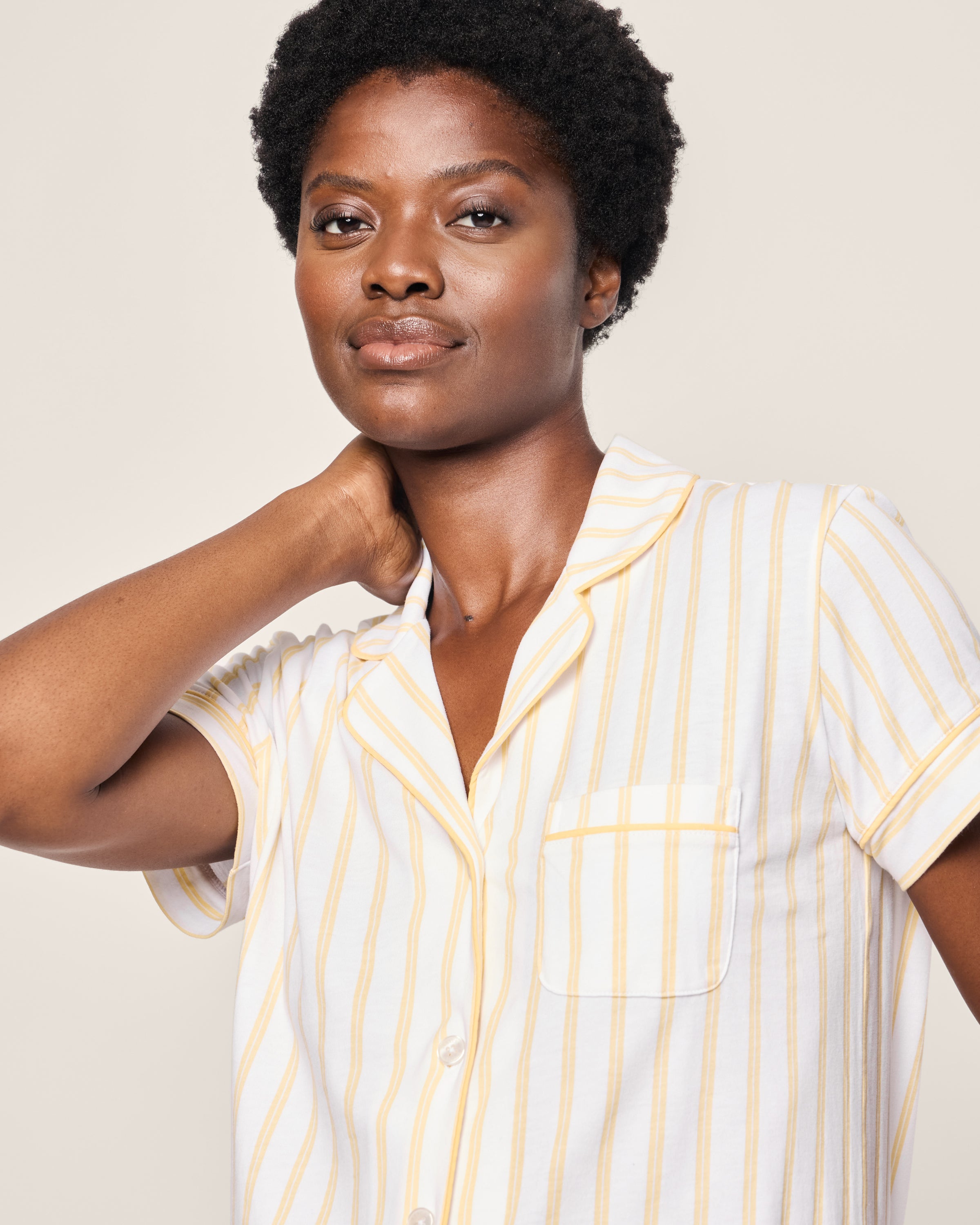 Wearing the Womens Pima Short Sleeve Short Set in Sunny Stripe by Petite Plume, a person with an afro hairstyle touches their neck and gazes slightly past the camera with a neutral expression.