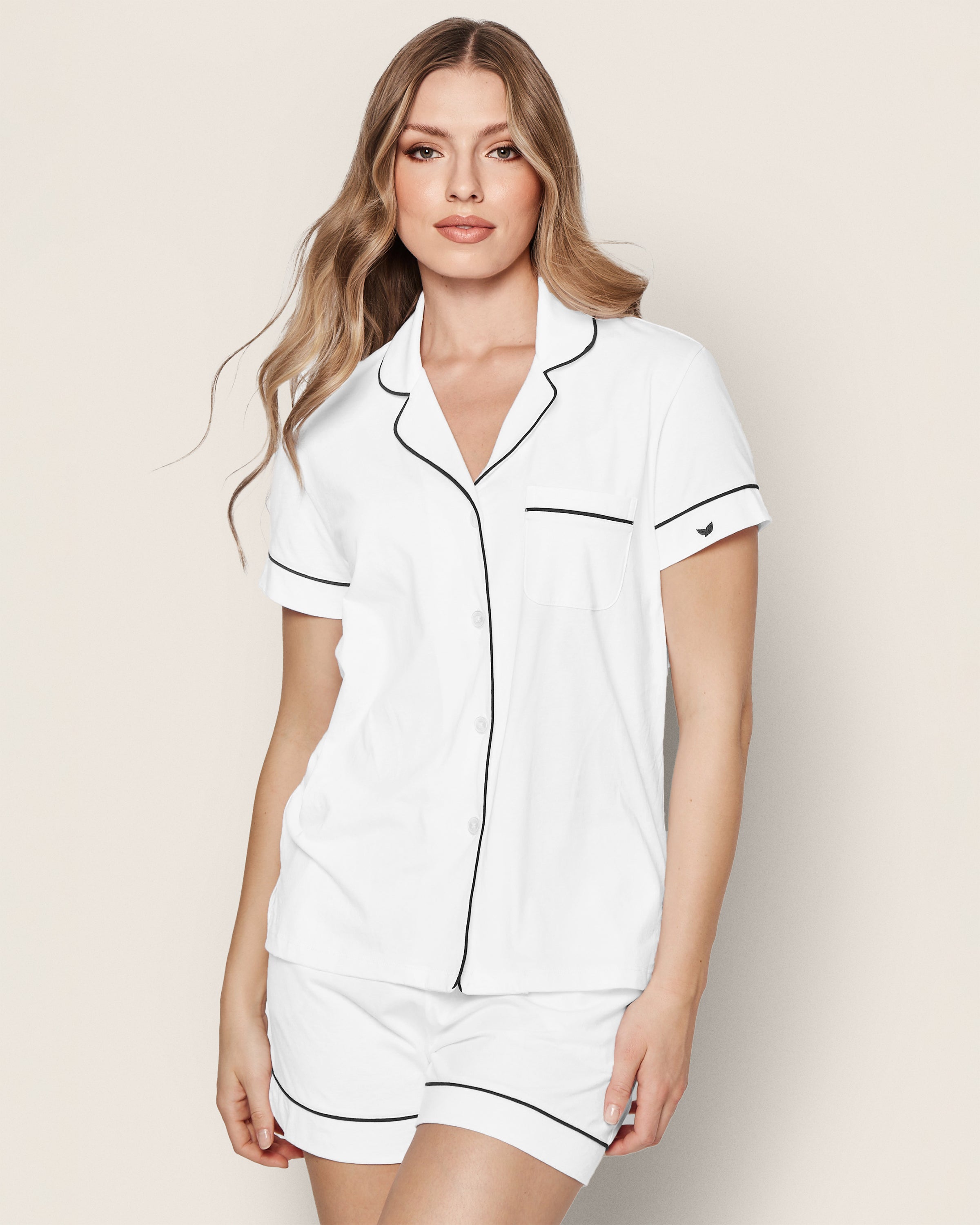 A woman with long hair models Petite Plumes Womens Pima Pajama Short Set in white with navy piping, featuring a short-sleeved, collared top with buttons and a pocket. She poses gracefully against a neutral backdrop, showcasing comfort and style.