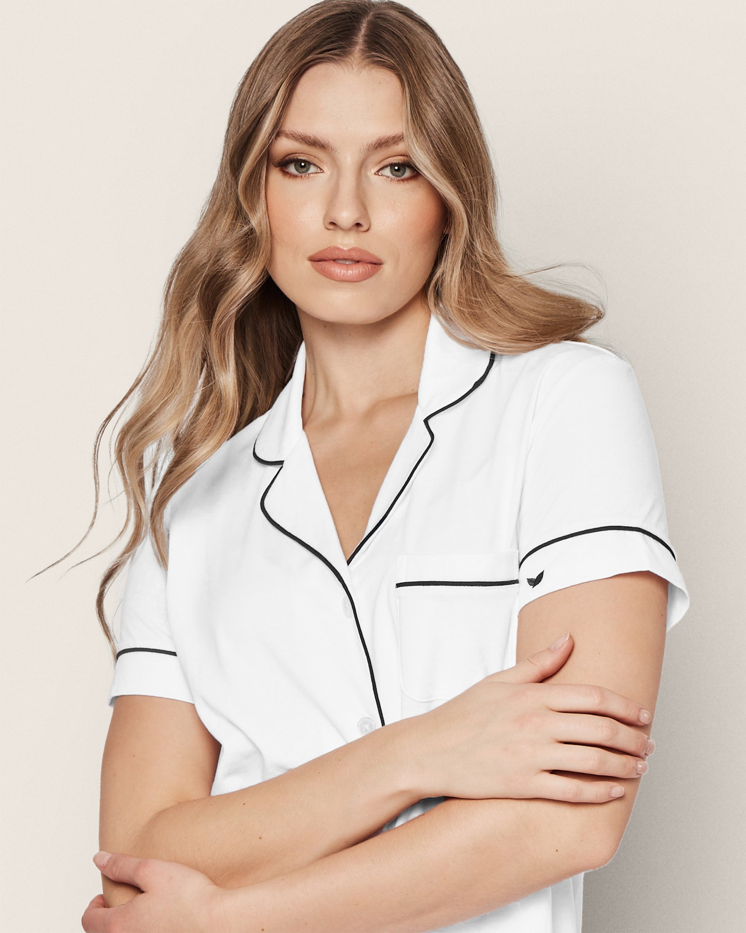 A person with long, wavy blonde hair wears a Womens Pima Pajama Short Set in White with Navy Piping by Petite Plume. They have a neutral expression and face the camera against a plain, light background, enhancing the sense of understated luxury.