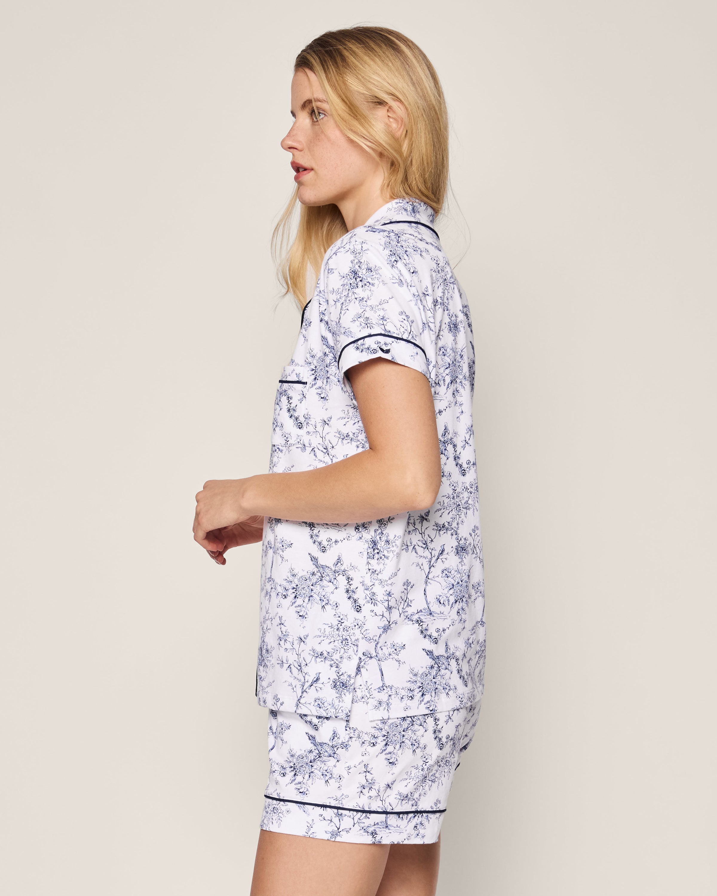 A person with long hair stands in profile wearing Petite Plumes Womens Pima Short Sleeve Short Set in Timeless Toile, showcasing a classic blue floral pattern on white. The image radiates botanical elegance against a neutral gray backdrop.