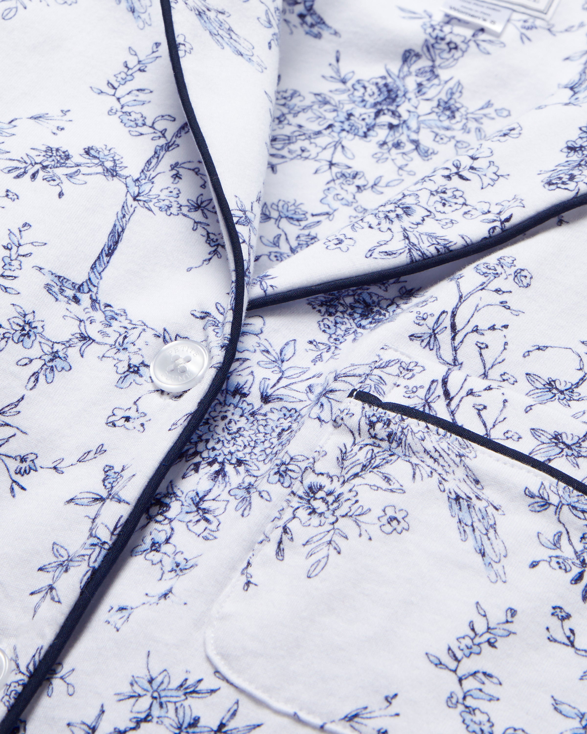 A close-up of the Womens Pima Short Sleeve Short Set in Timeless Toile by Petite Plume shows white fabric with delicate blue florals, dark piping on edges, a visible clear button, and part of a pocket, reflecting luxurious PJ elegance.